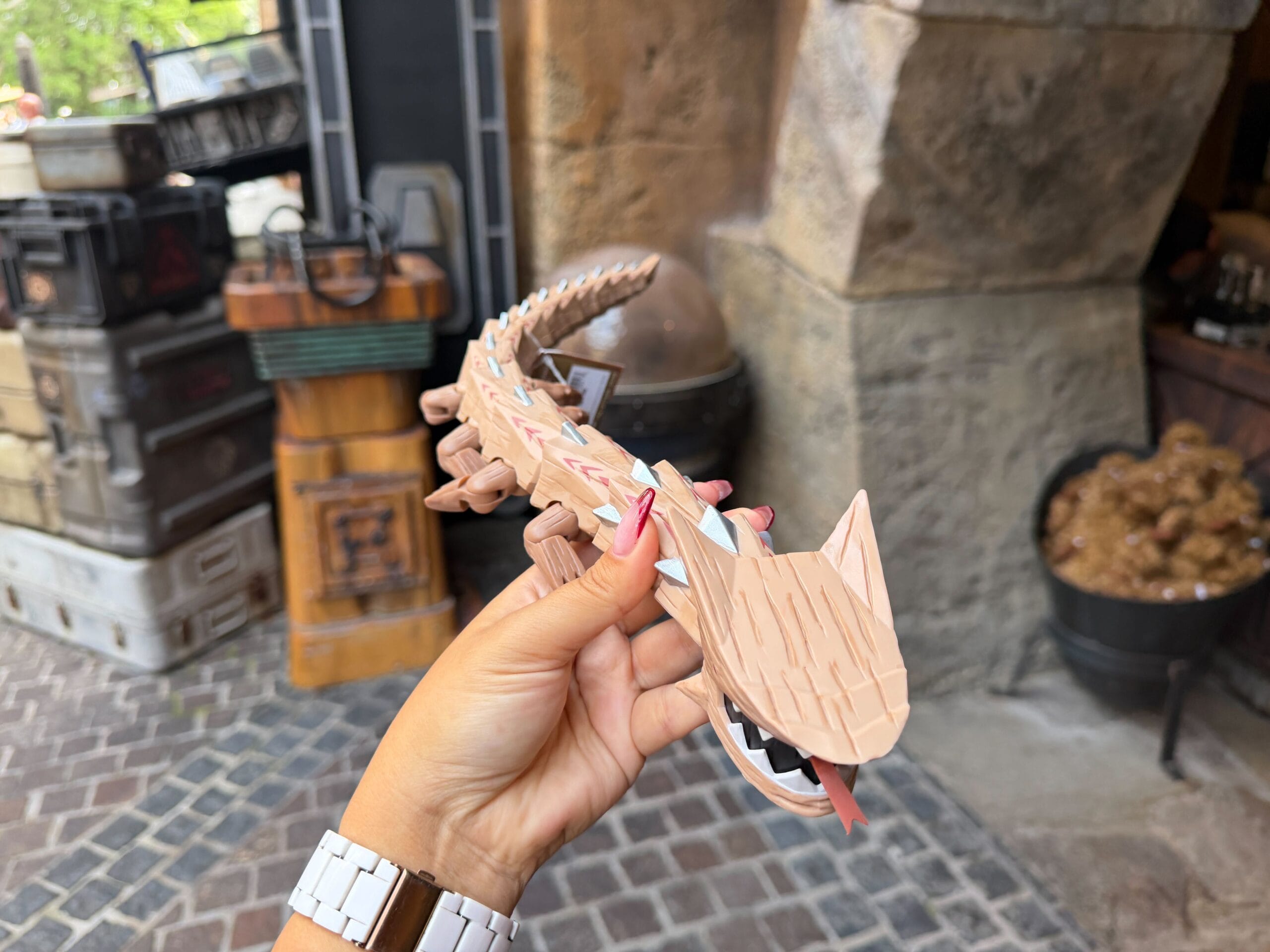 A hand holds a wooden Krayt Dragon toy with an open mouth at Galaxy’s Edge, the market bustling like those in Disney theme parks.