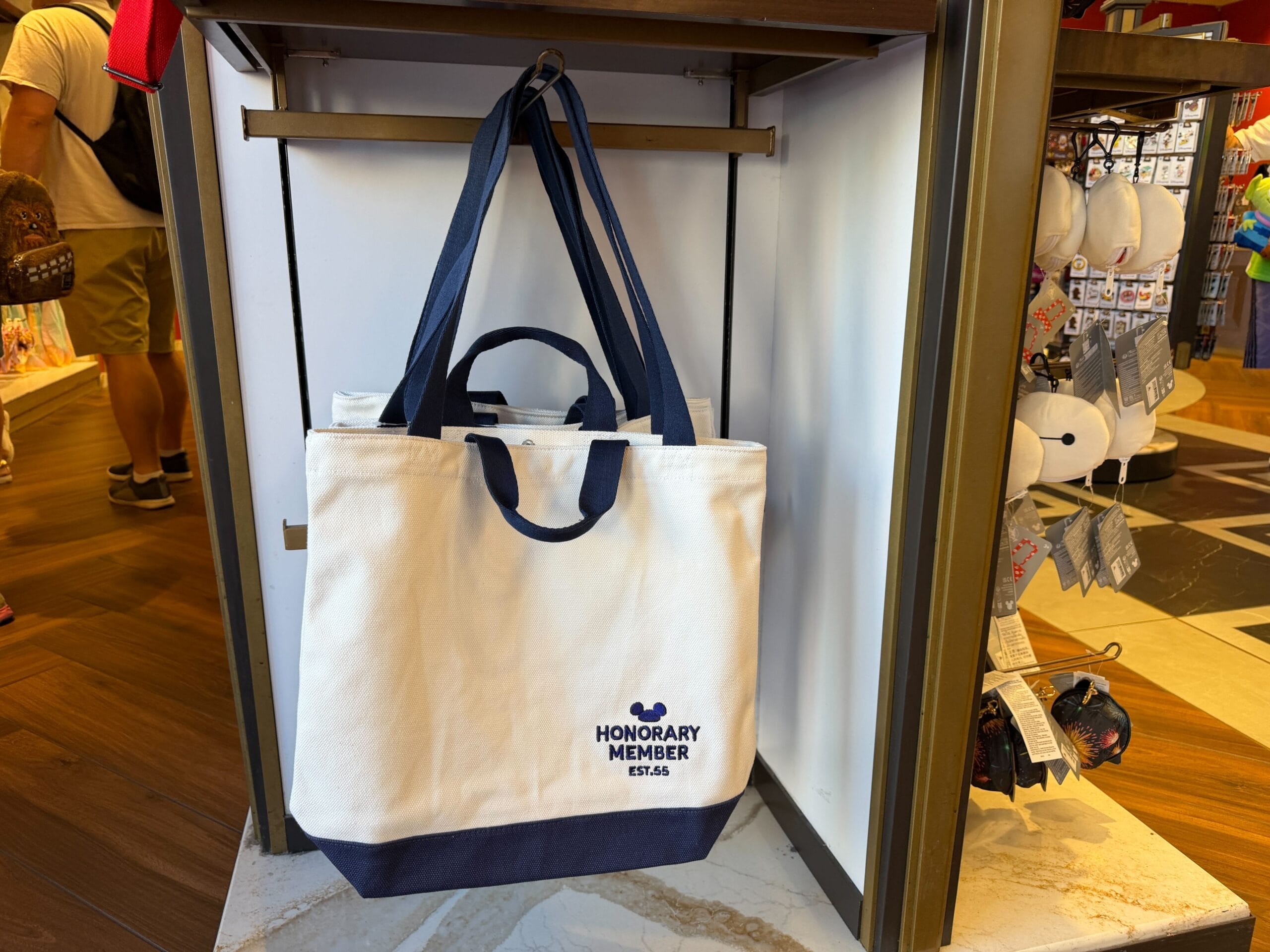 A white and navy tote bag with "Honorary Member" text sits on a store shelf, reminiscent of Disney Parks merchandise displays.