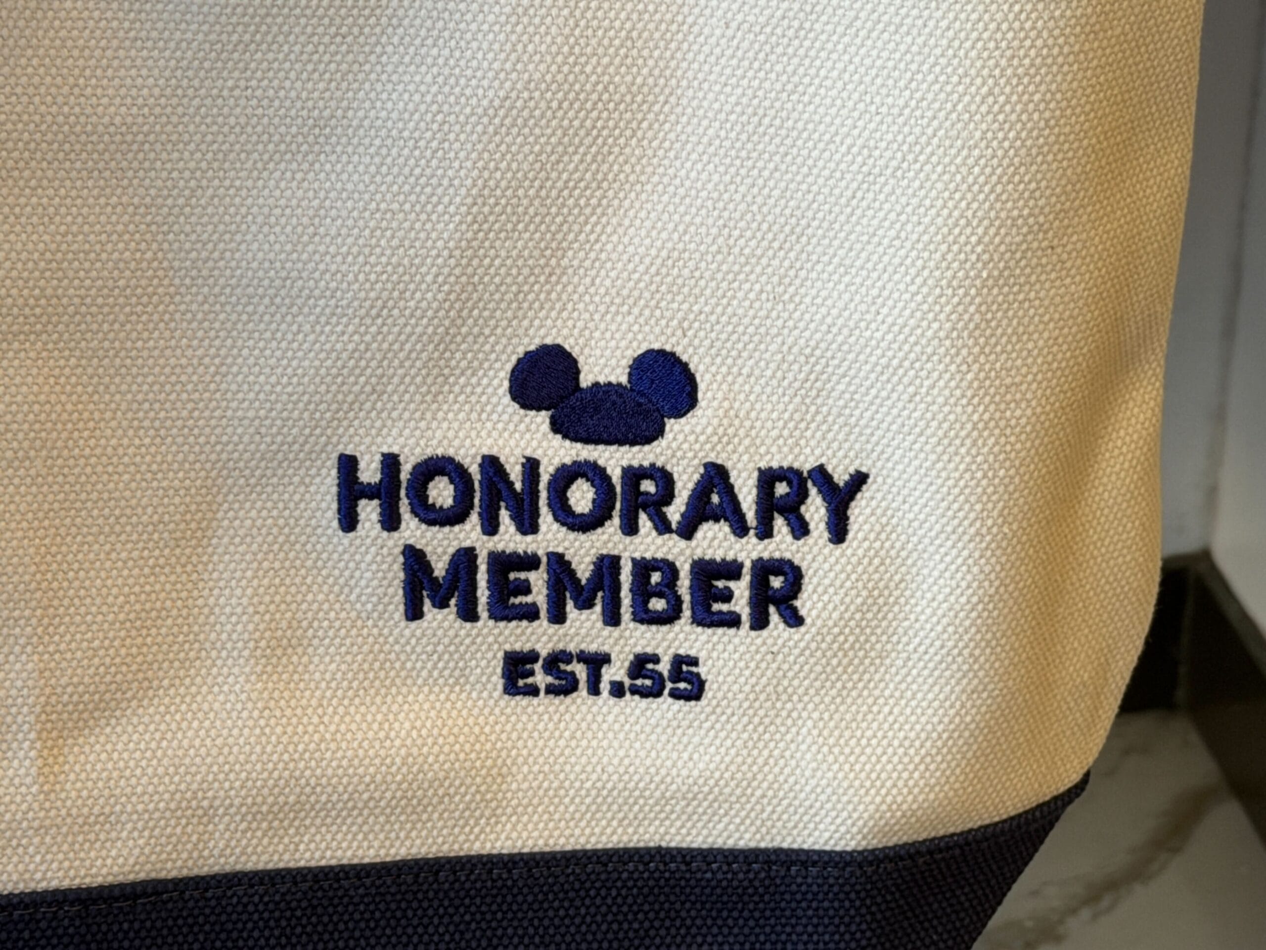 A fabric bag features "HONORARY MEMBER EST. 55" embroidered with a blue Mickey head icon above, evoking classic Disney Parks style.