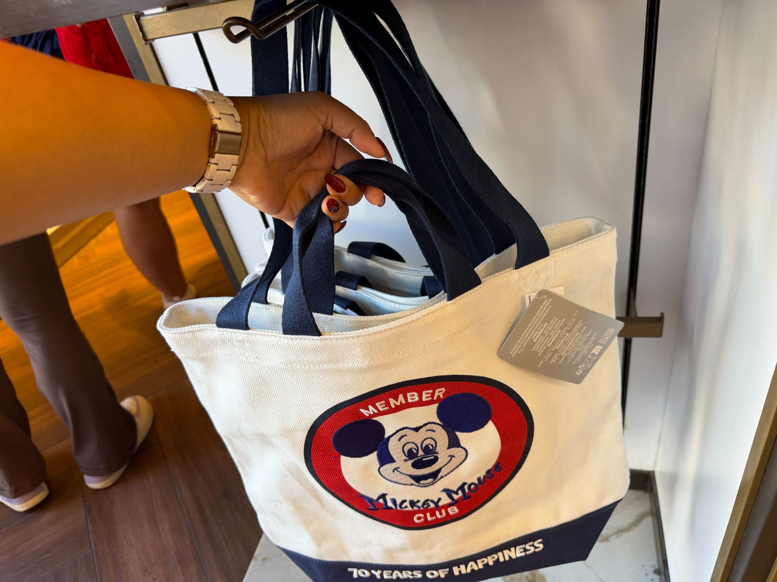 A person at a Disney park holds a tote bag featuring the Mickey Mouse Club logo and “70 Years of Happiness” in bold text.