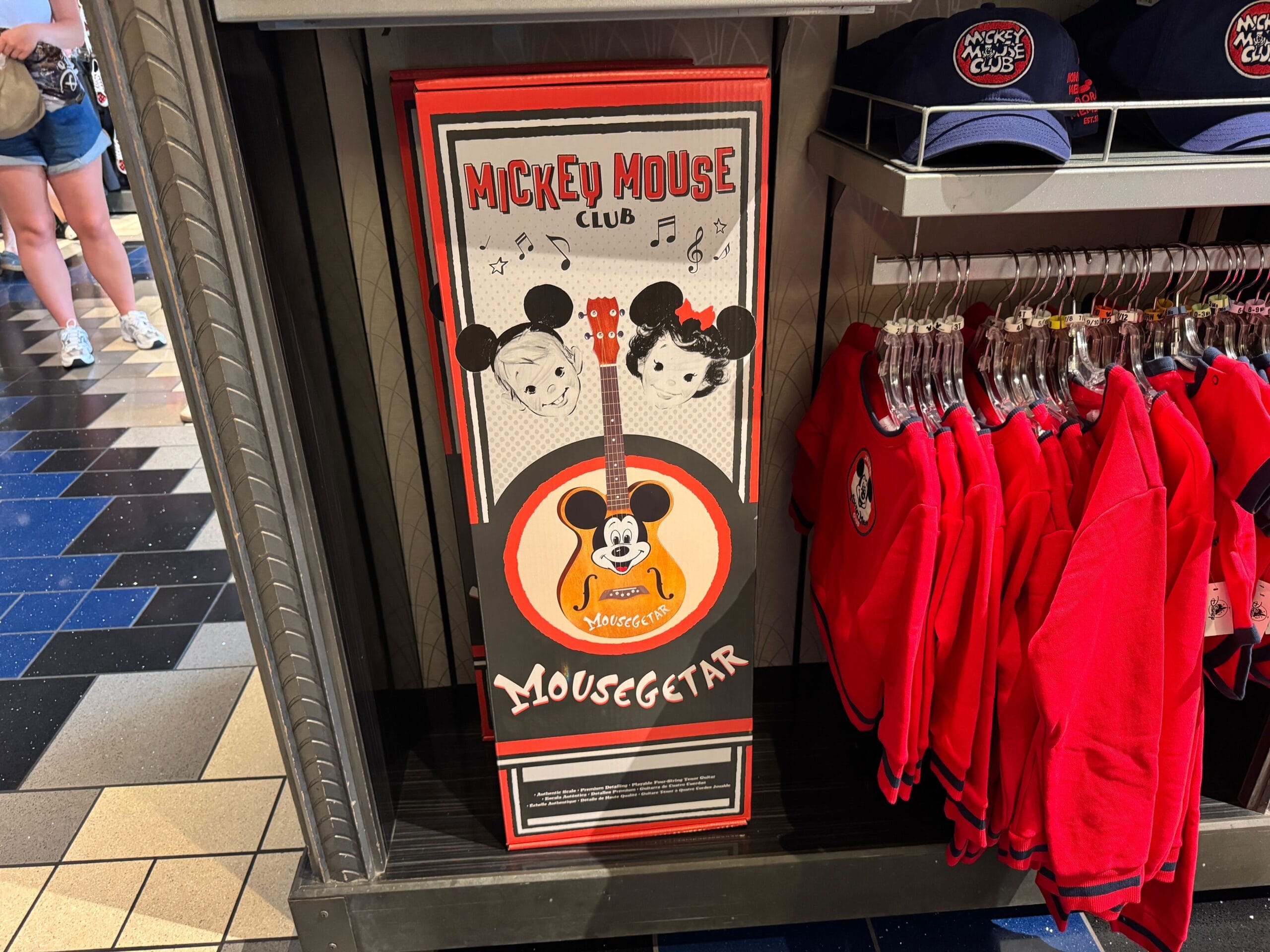 A boxed "Mousegetar" guitar featuring Mickey Mouse art is on display beside red apparel in a Disney park merchandise shop.