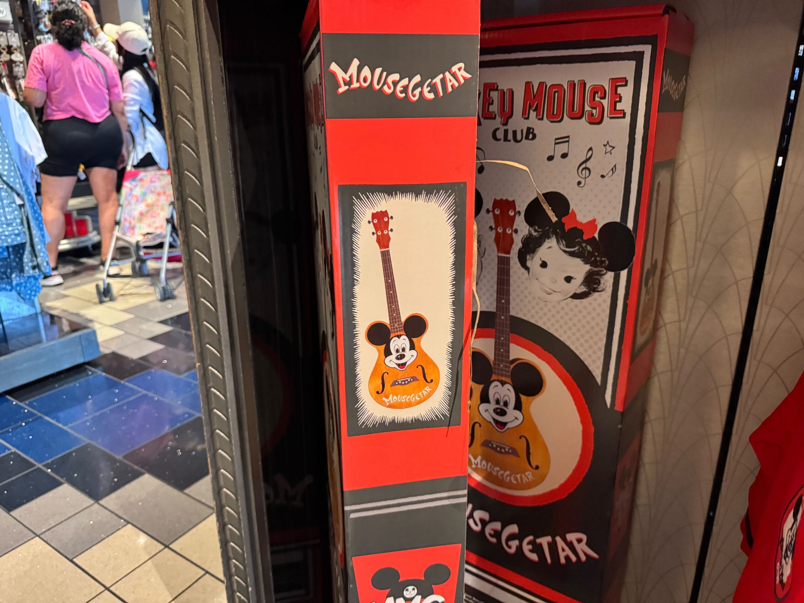 A store display box shows a whimsical guitar featuring Mickey Mouse’s face, called "Mousegetar"—classic Disney park souvenir style.
