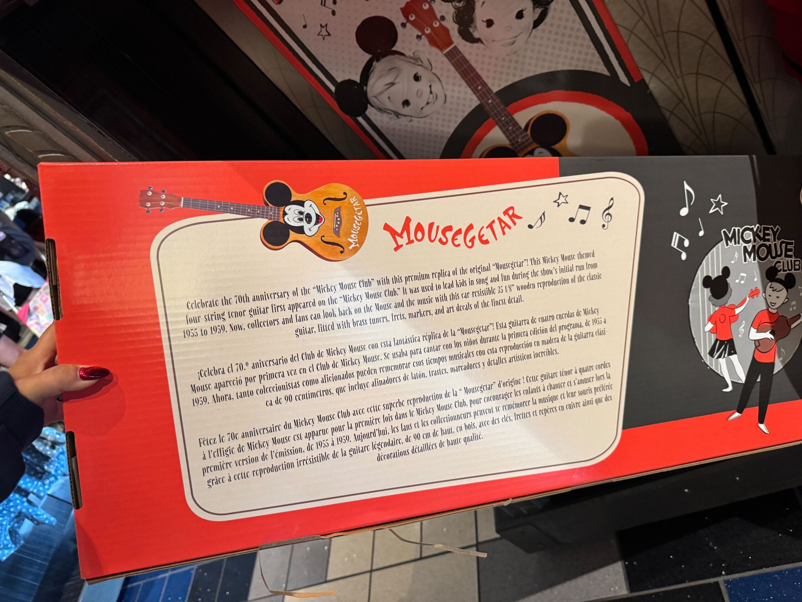 A person holds a red Disney Mousegetar box featuring Mickey Mouse and Mickey Mouse Club guitar details, reminiscent of park merchandise.