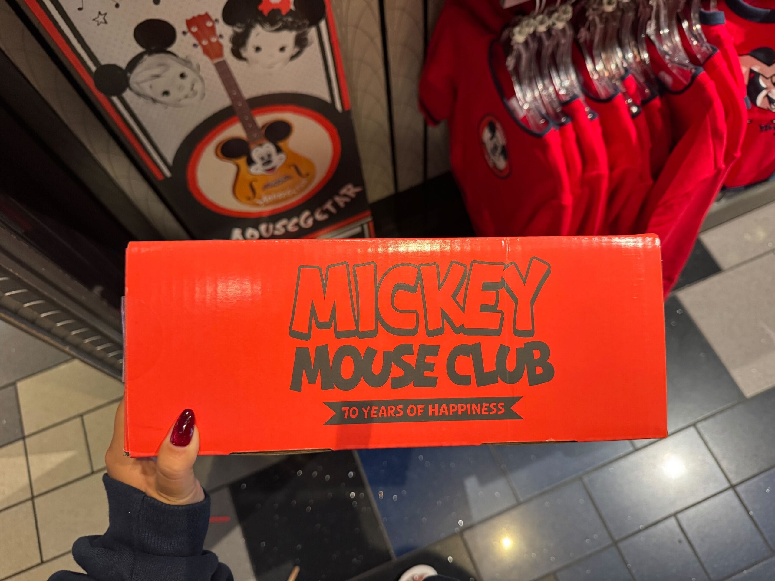 A hand holds a red "Mickey Mouse Club, 70 Years of Happiness" box in a Disney park shop filled with Mickey-themed merchandise.