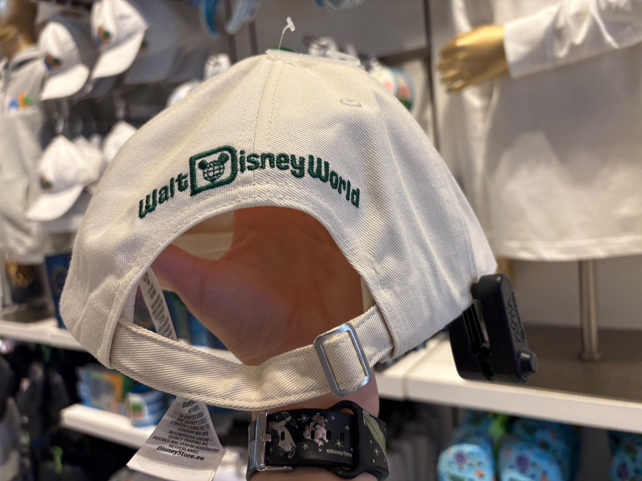 A hand holds a white Walt Disney World cap with green embroidered text, displayed in a shop near Cinderella Castle at Magic Kingdom.