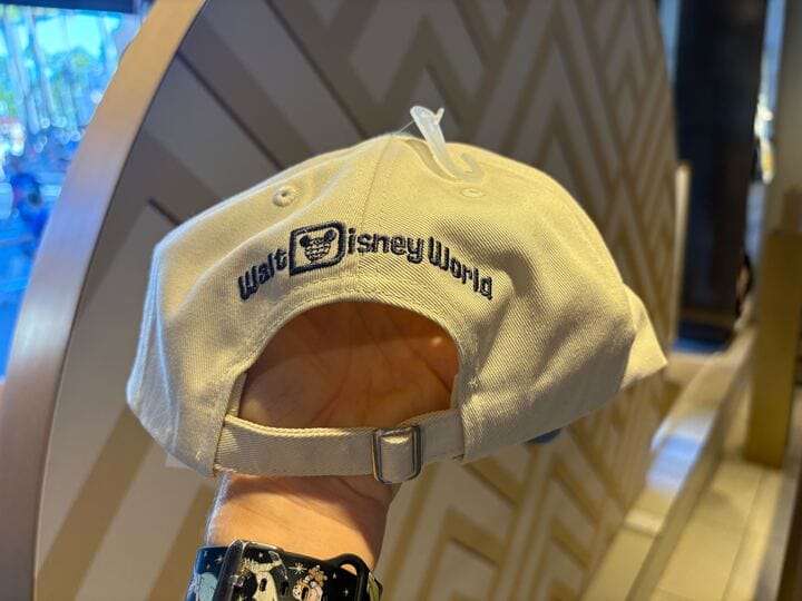 A hand displays a beige Walt Disney World cap with dark blue logo embroidery, reminiscent of classic park merchandise styles.