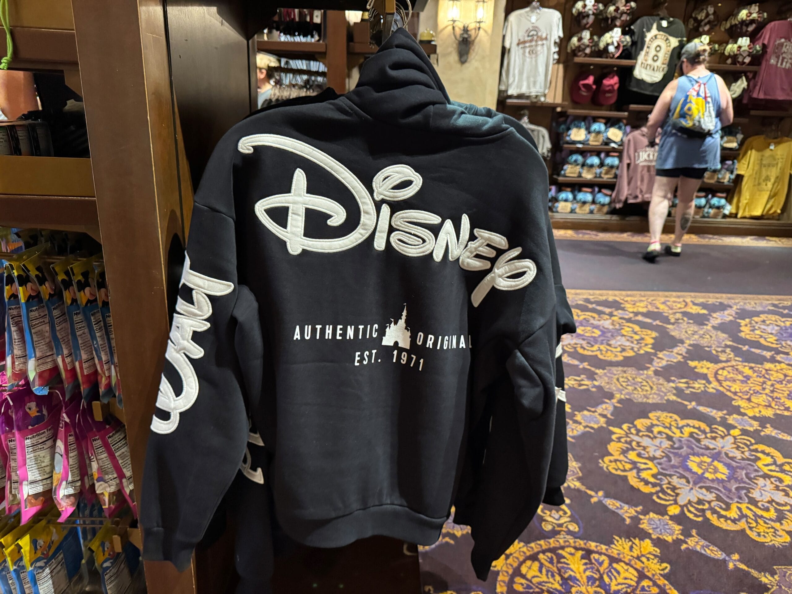 A black Disney hoodie with white lettering is showcased in a park souvenir shop with purple carpet, surrounded by shoppers.