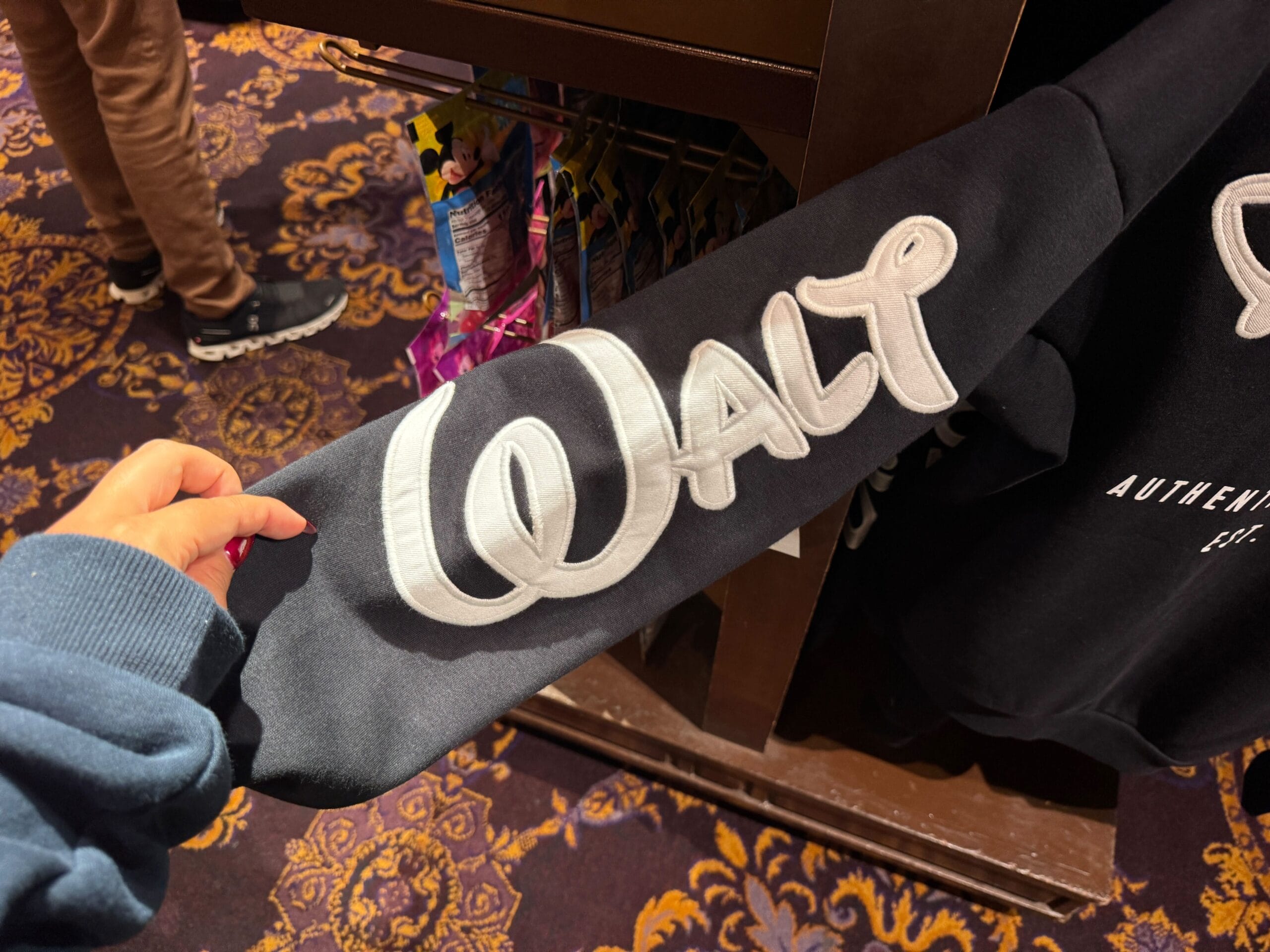 A hand holds a black sweatshirt sleeve bearing “Walt” in white script, reminiscent of classic Disney Parks merchandise style.
