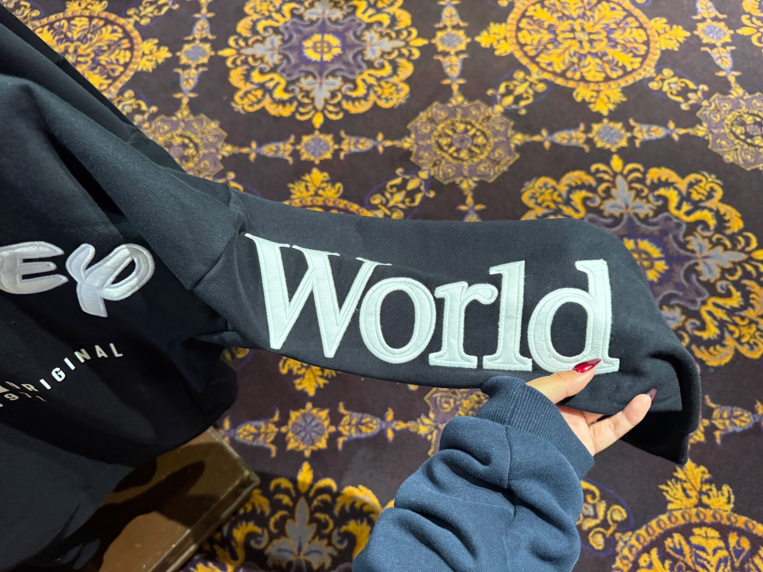 A person holds a black sleeve with "World" embroidered, above a patterned carpet reminiscent of Disney park hotel carpeting.
