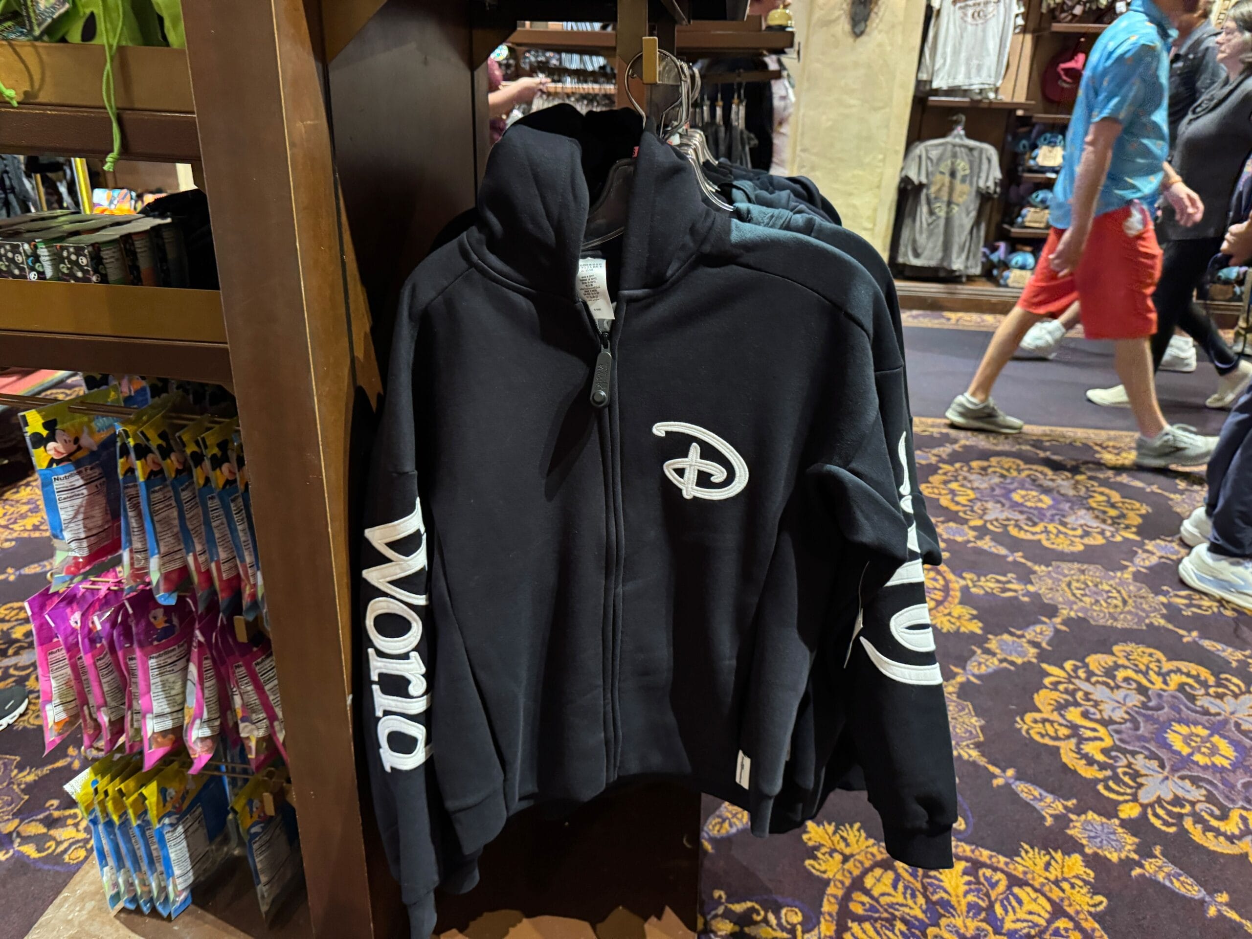 A black zip-up jacket with a Disney "D" logo and "World" on the sleeves, seen hanging in a Walt Disney Parks store display.