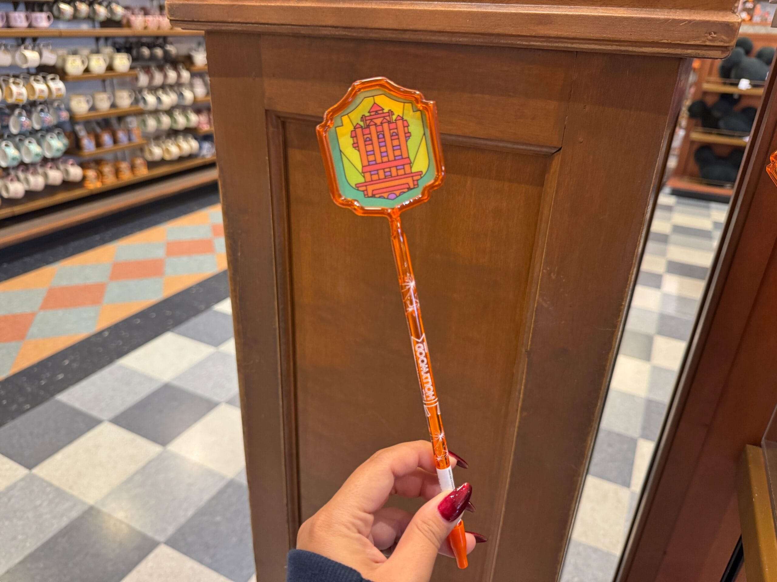 A hand holding a Hollywood Studios pen with a vibrant topper in front of a wooden display inside a Walt Disney World store.