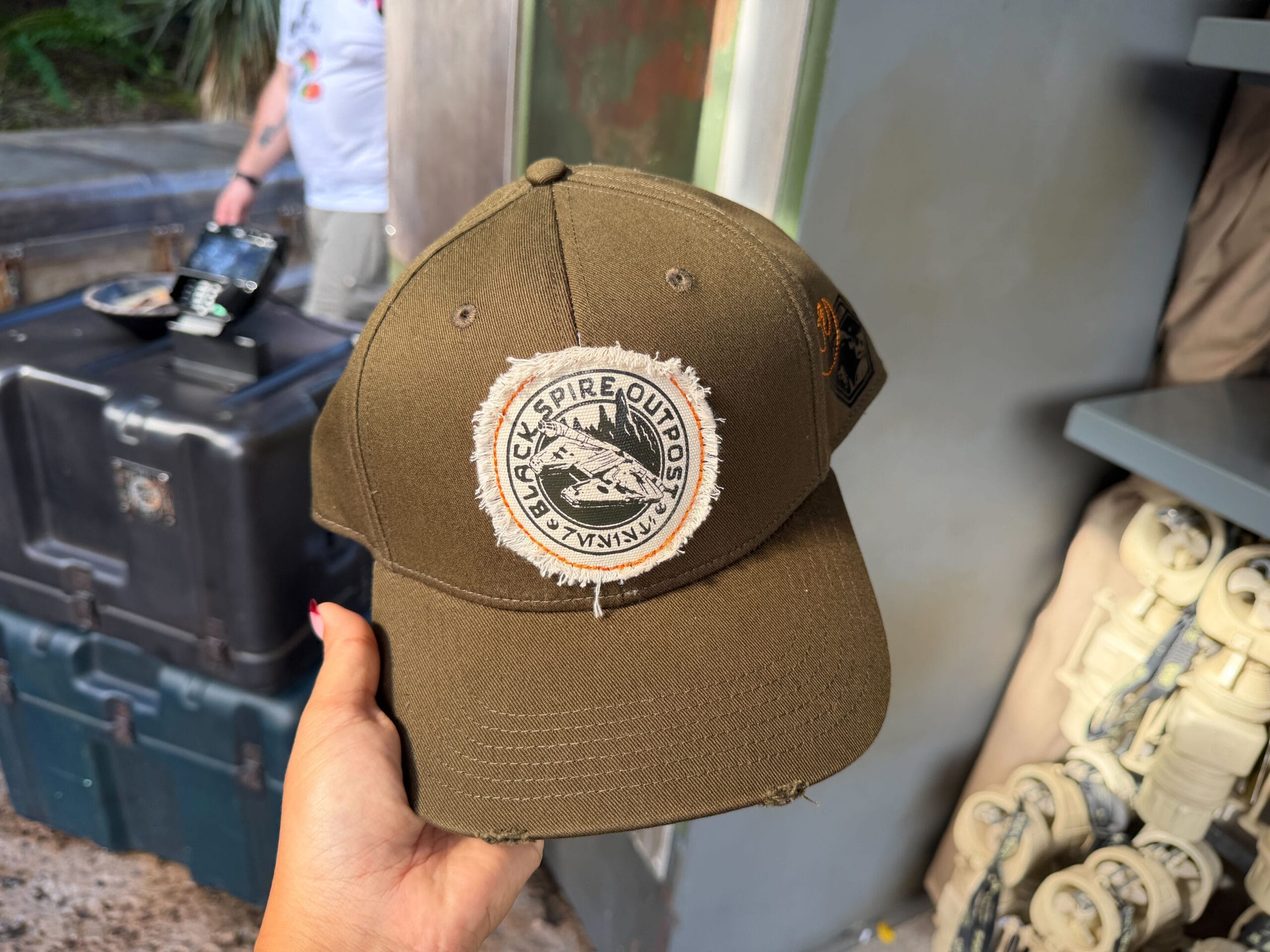 A hand holds an olive-green "Black Spire Outpost" hat, a Star Wars: Galaxy’s Edge souvenir, in front of a themed park shelf display.