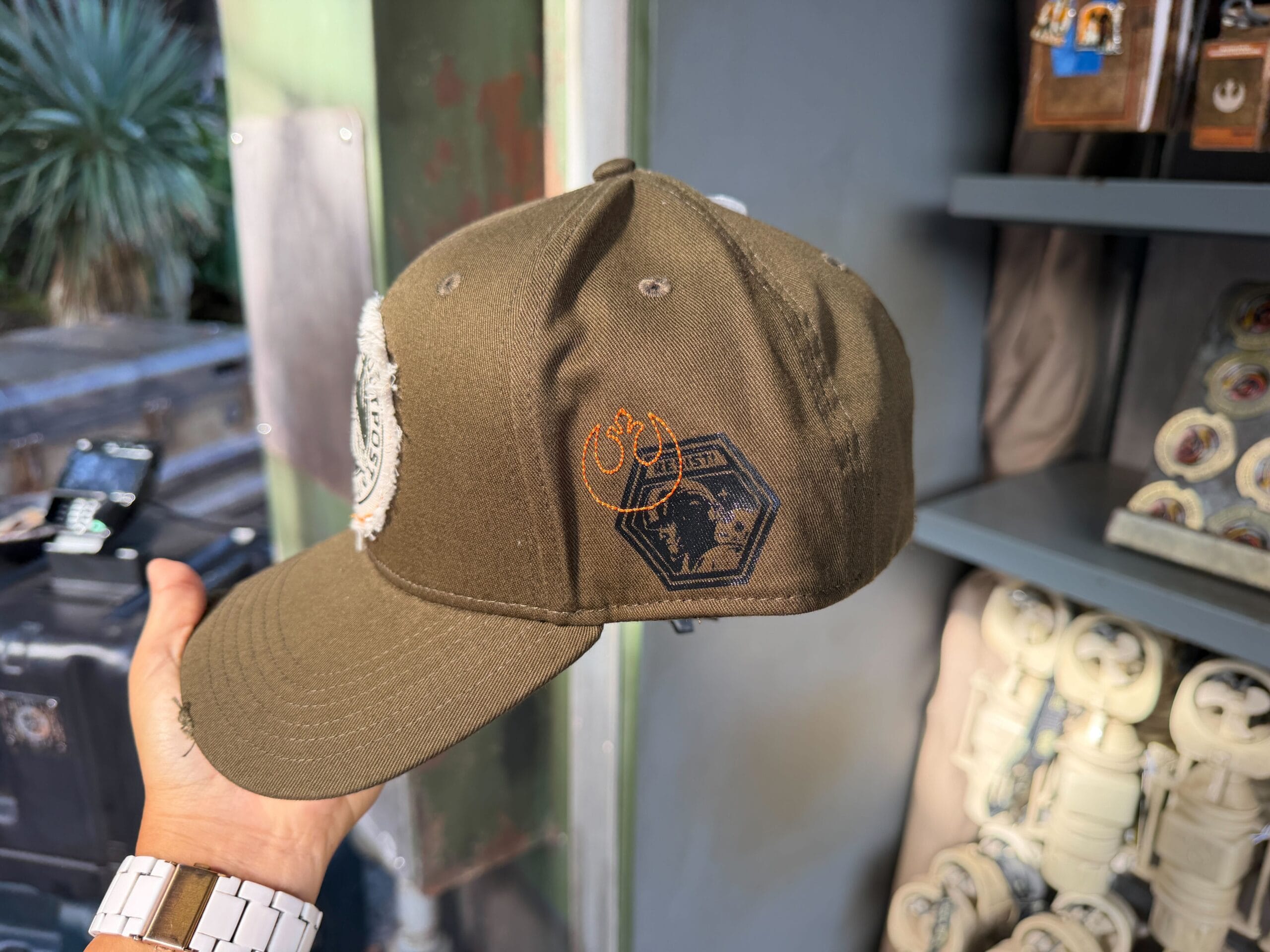 A hand holds an olive green Star Wars Resistance cap, similar to merchandise at Disney parks, near a themed display shelf.