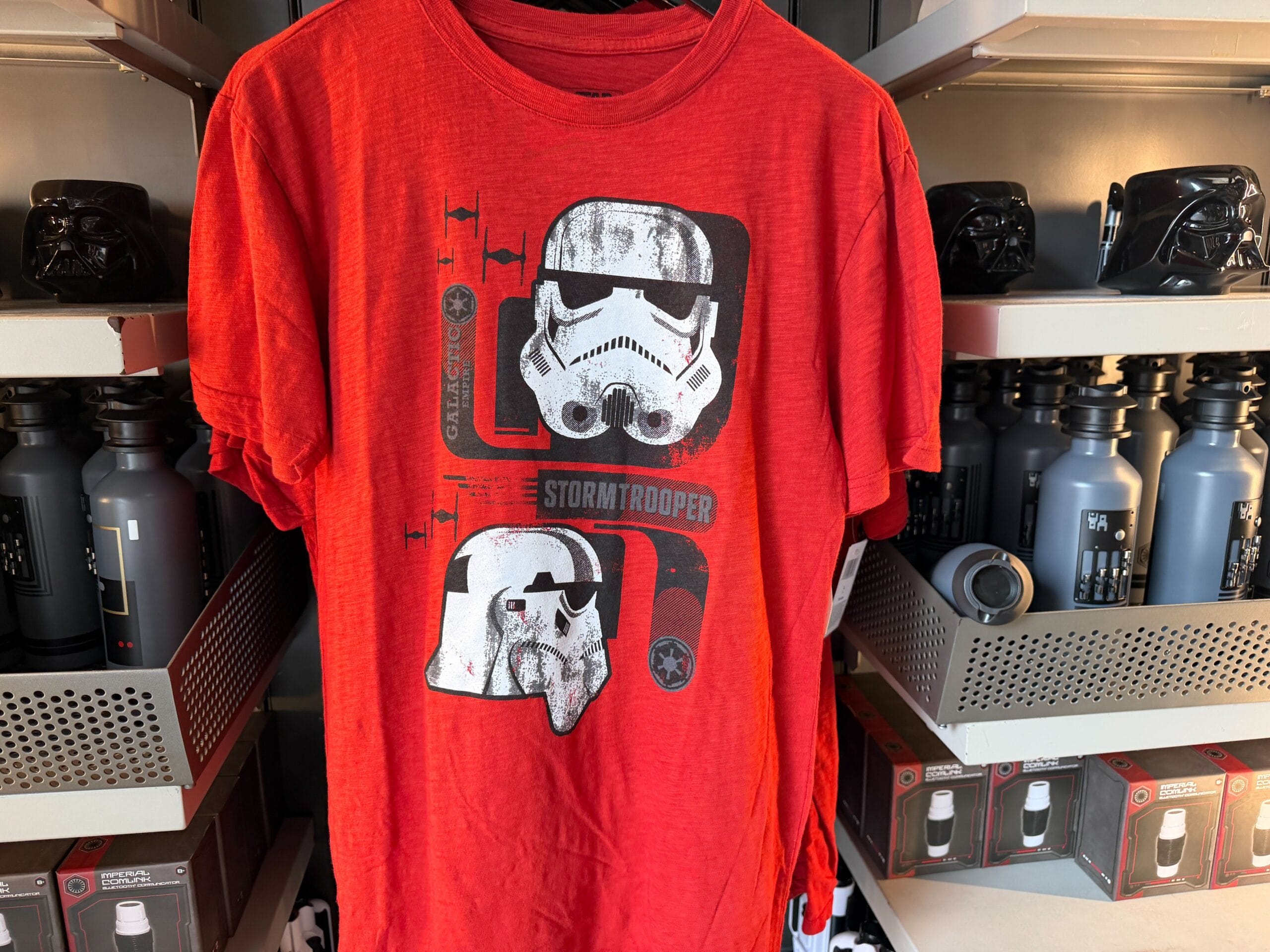 Red T-shirt with two white stormtrooper helmets and "Stormtrooper" text, displayed among black water bottles at a Disney park.