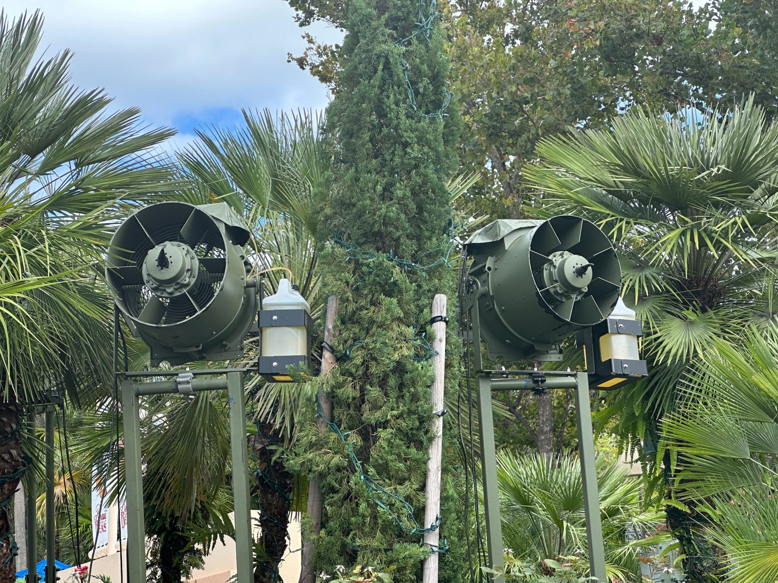 Two large green fans on metal stands with special effects equipment, set outdoors among lush palms and greenery, Disney park style.