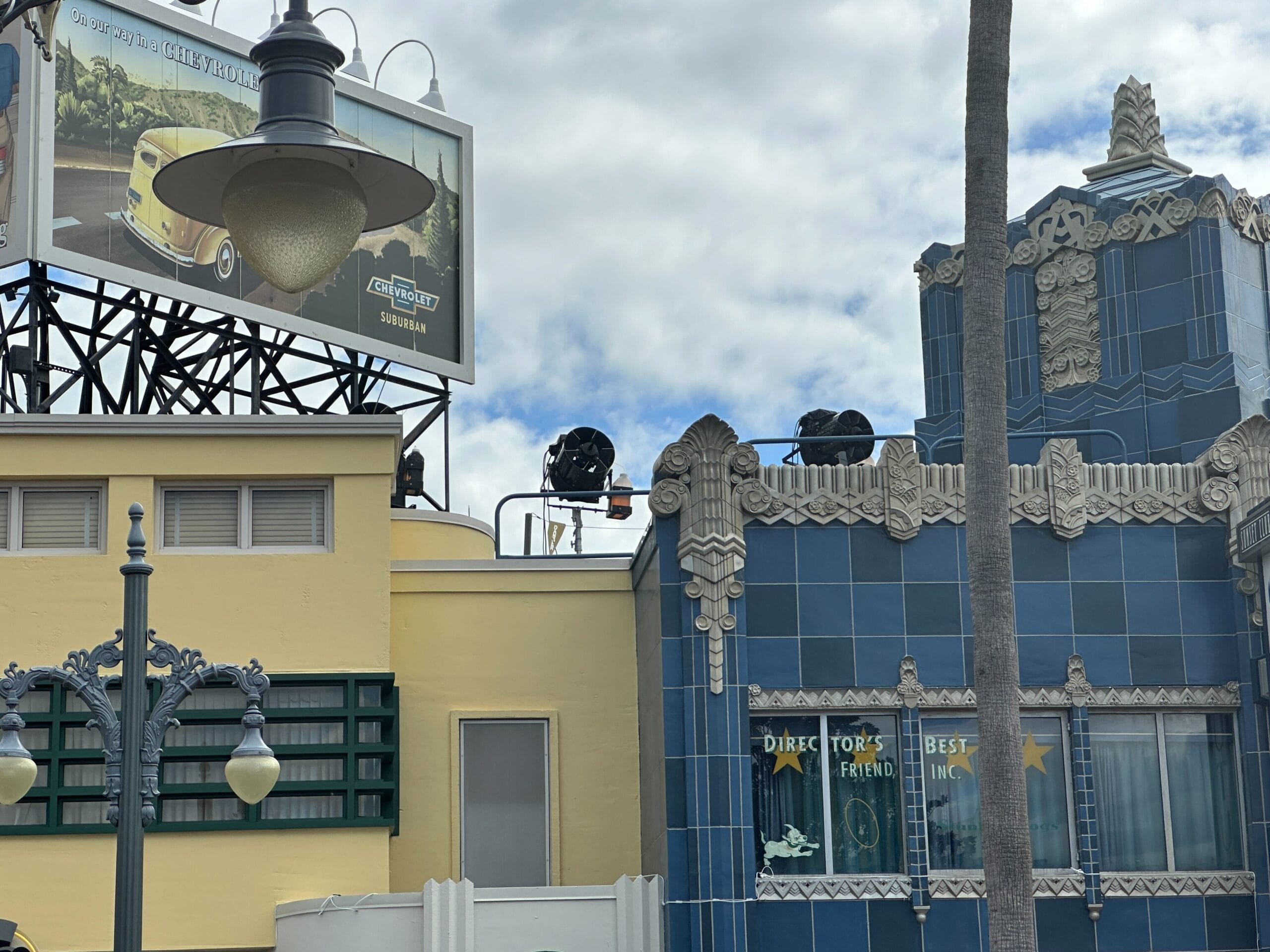 Hollywood Studios art deco building with blue tiles, palm tree, vintage lamps, and yellow car billboard beneath cloudy skies.