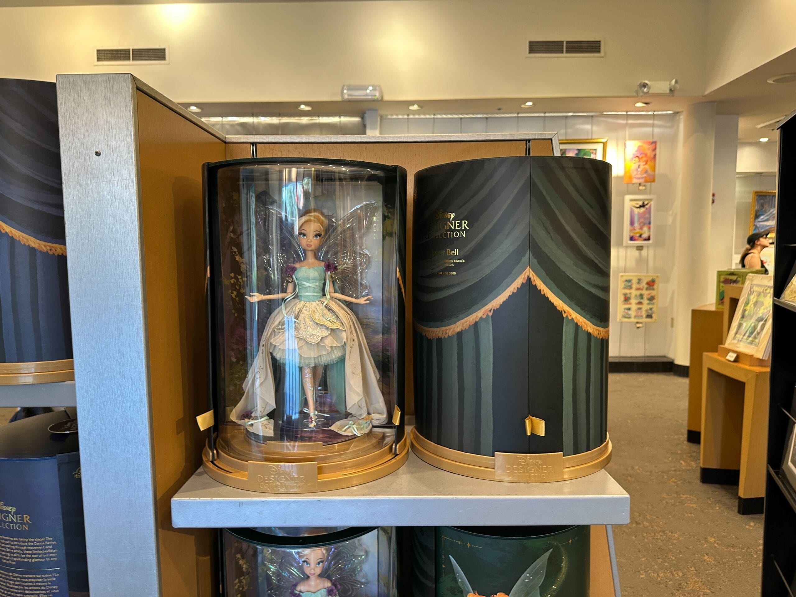 A Designer Tinker Bell doll in a display case sits on a Disney Parks store shelf beside its ornate cylindrical packaging.
