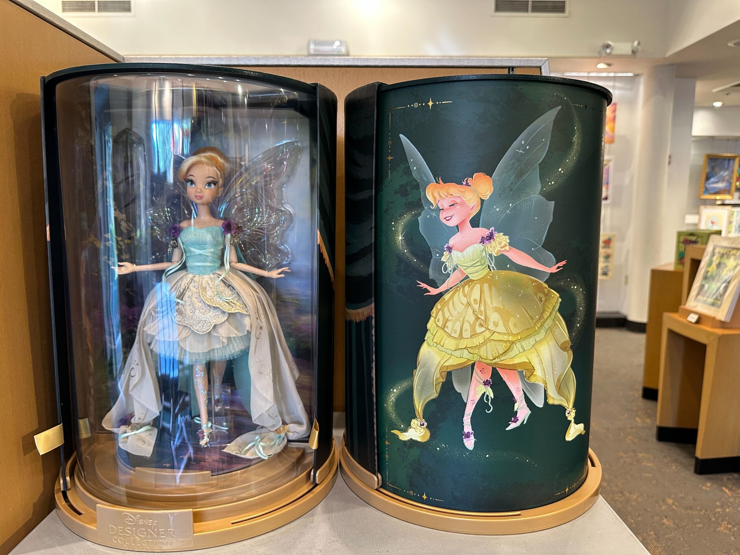 A Designer Tinker Bell doll in a display case stands next to an illustrated Tinker Bell, both against a Disneyland-inspired green backdrop.