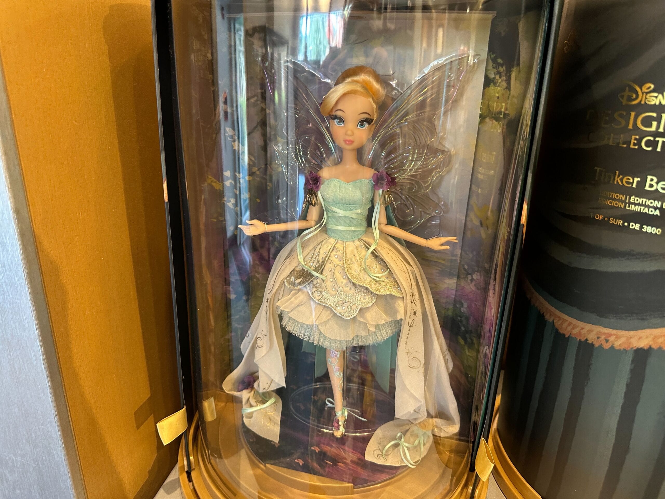 A collectible Designer Tinker Bell doll in a display case at a Disney park, dressed in elaborate attire with wings and fine details.