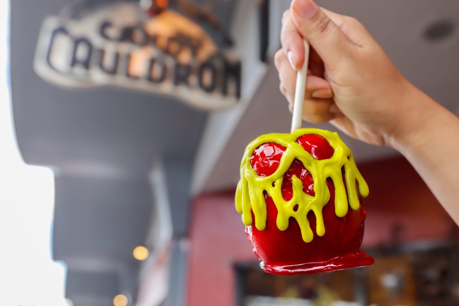 A hand holds a red candy apple with green icing in front of the Cauldron shop at Disney—no castle in sight this time!