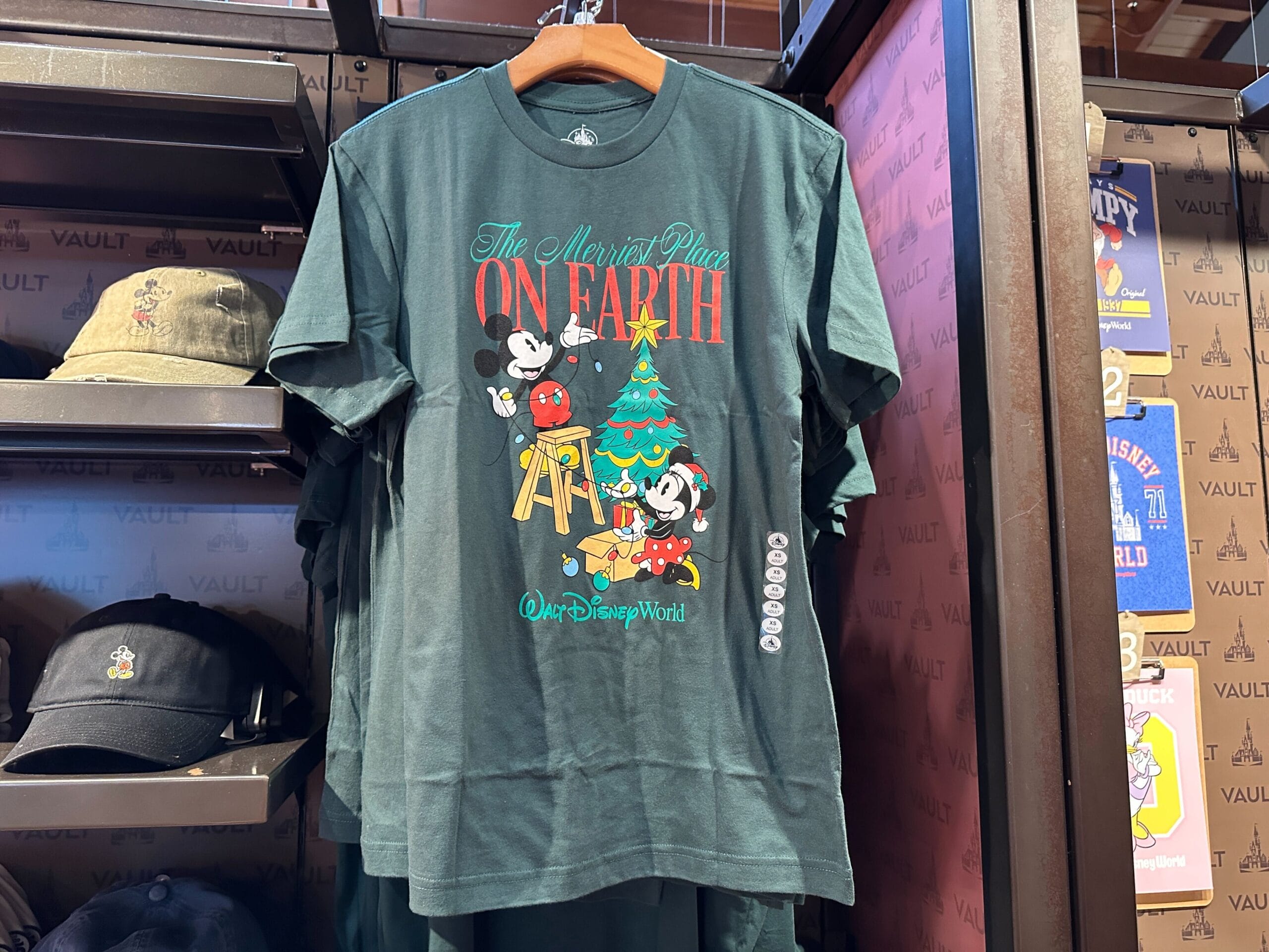 Green holiday tee with Mickey and Minnie decorating a Christmas tree at Disney World, labeled "The Merriest Place on Earth.