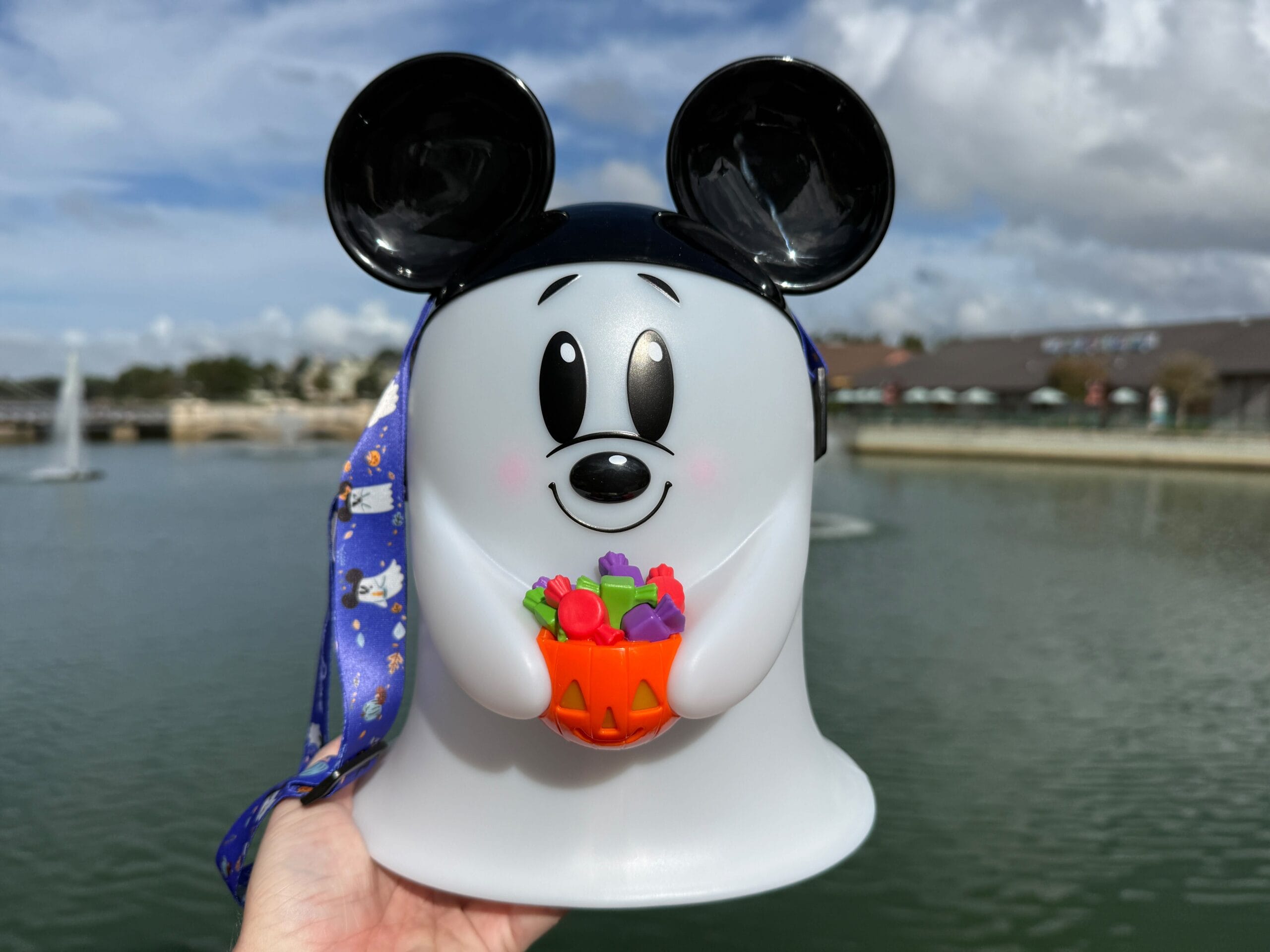 A hand holds a Mickey Mouse Ghost Popcorn Bucket filled with candy beside a lake, clouds above, at a Disney park—not a castle view.