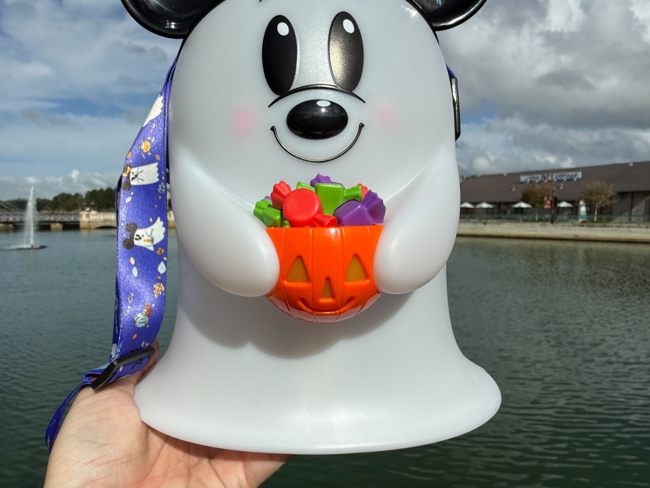 A hand displays a Ghost Popcorn Bucket with a pumpkin basket by a Disney World lake; buildings visible, no castle in this view.