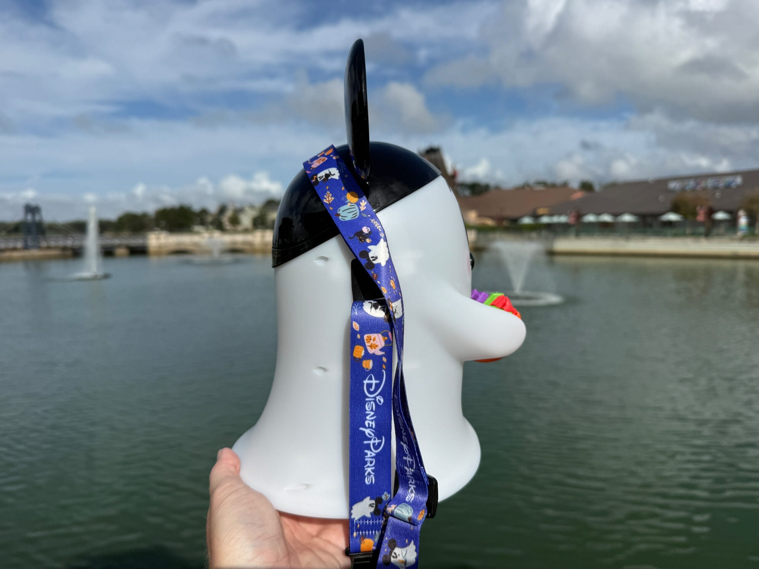 A hand holds a Donald Duck popcorn bucket with a Disney Parks lanyard at Disney World, next to a lake and water fountains.