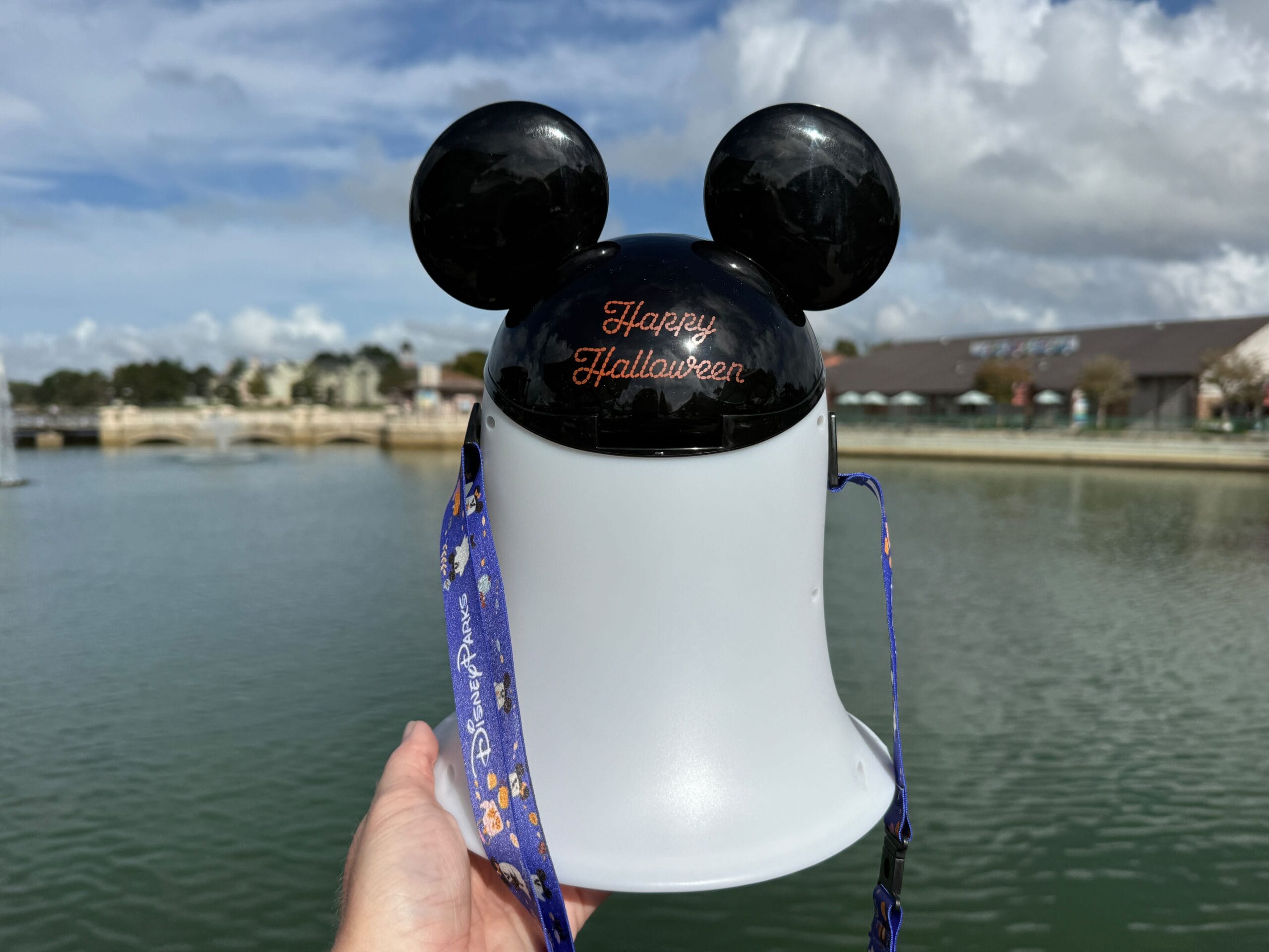 A hand holds a Mickey Mouse Ghost Popcorn Bucket by the water at a Disney park, with blue sky and distant buildings visible.
