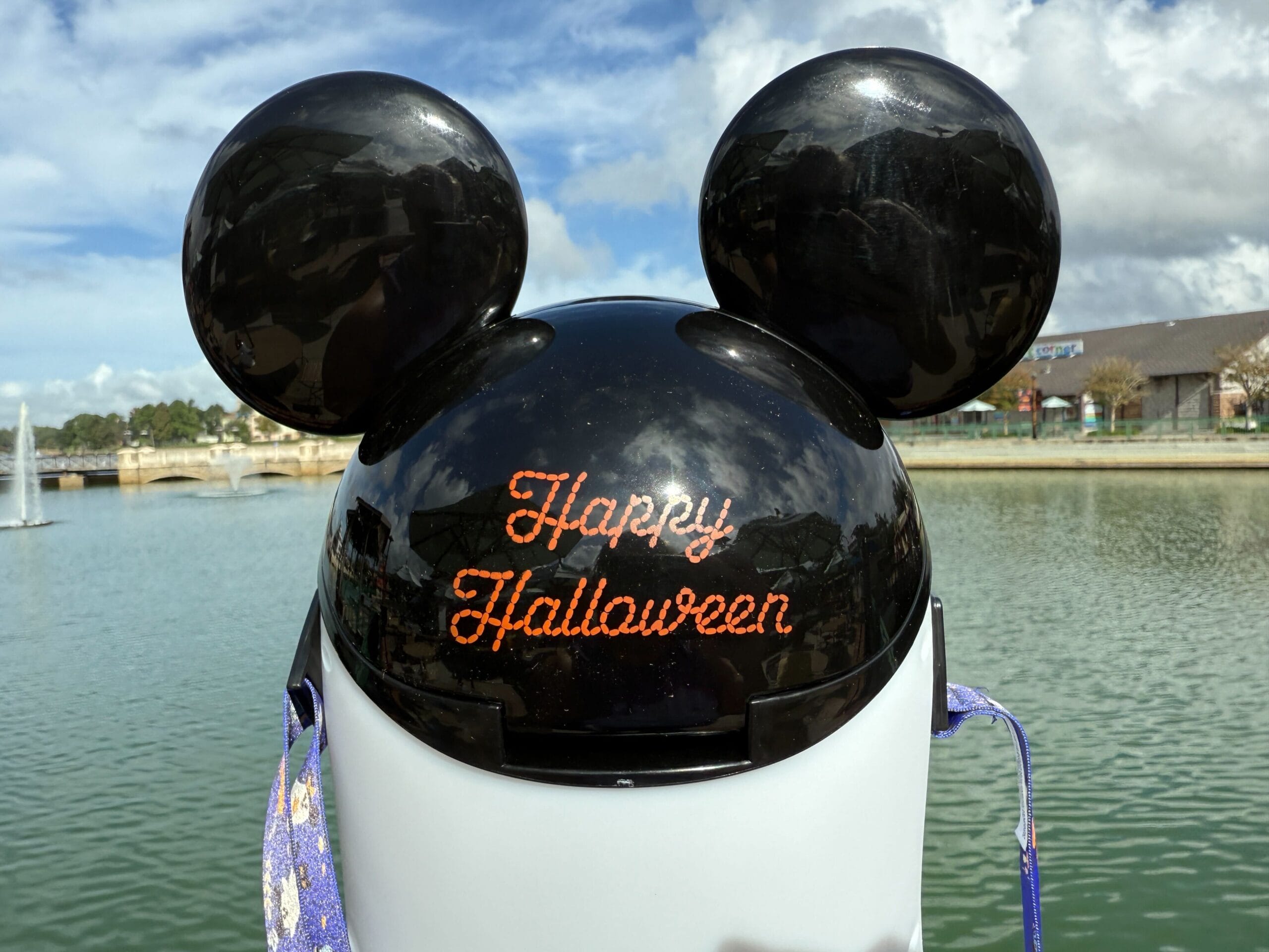 Mickey Mouse ear trash can with "Happy Halloween" text by a Disney World lake, sky partly cloudy—no castle in view to compare.