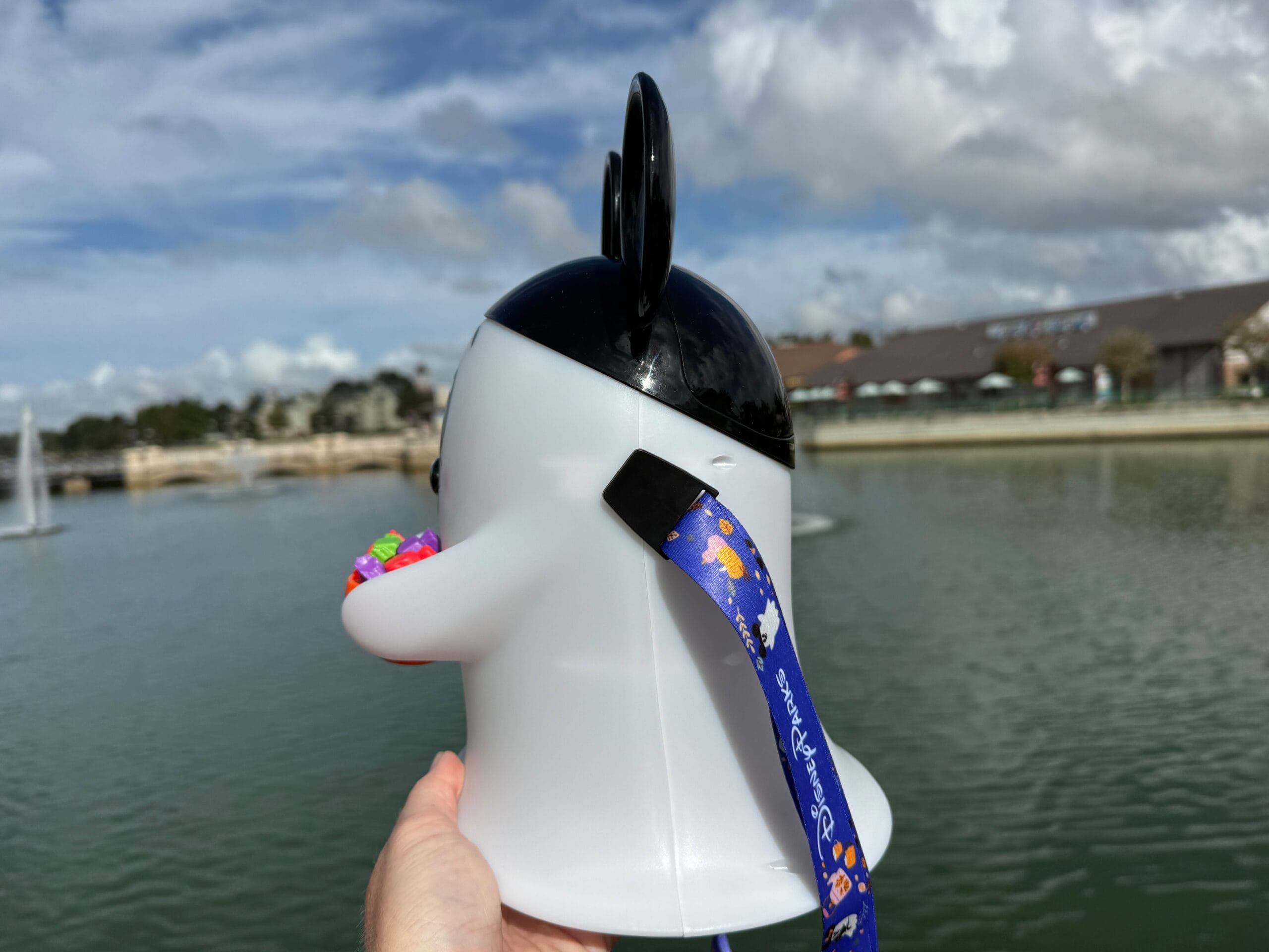 A hand holds a penguin-shaped Ghost Popcorn Bucket with a blue lanyard by a Disney park lake, sky dotted with clouds.