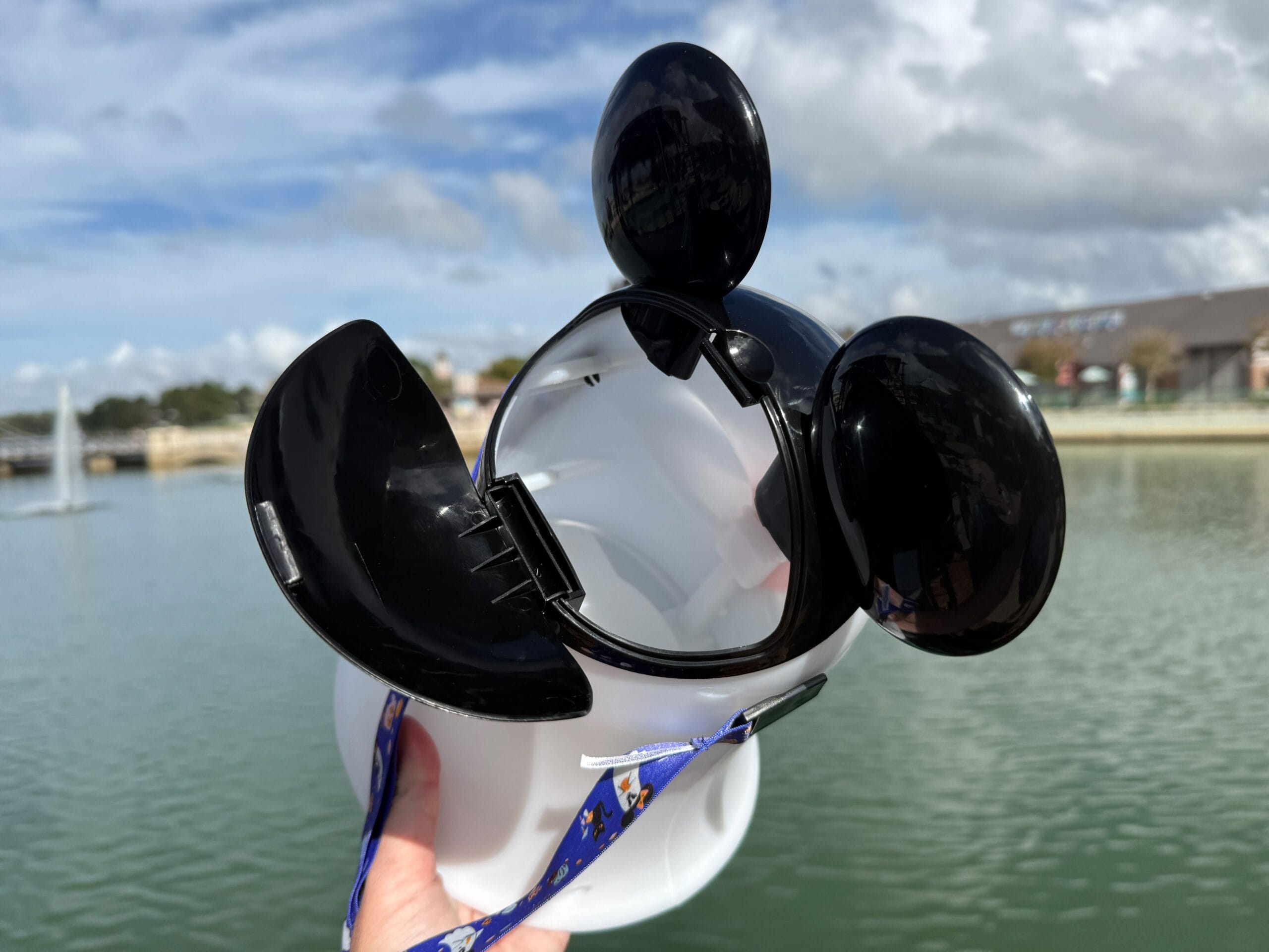 A hand holds an open Mickey Mouse Ghost Popcorn Bucket by a lake at Disney World on a cloudy day, no castle in view.