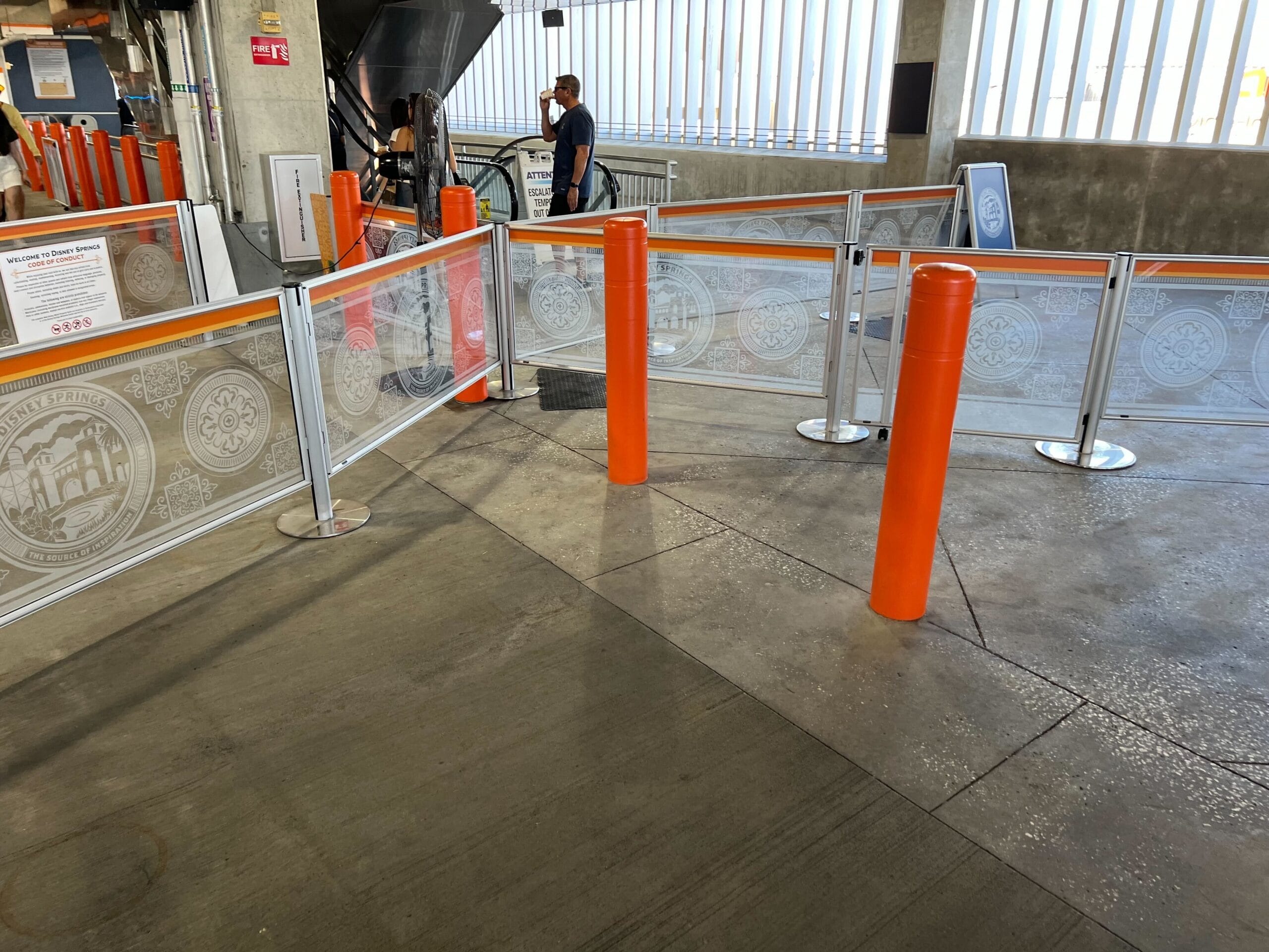 Orange queue posts and metal barriers line a concrete indoor space, resembling a Disney queue area, with one person in the distance.