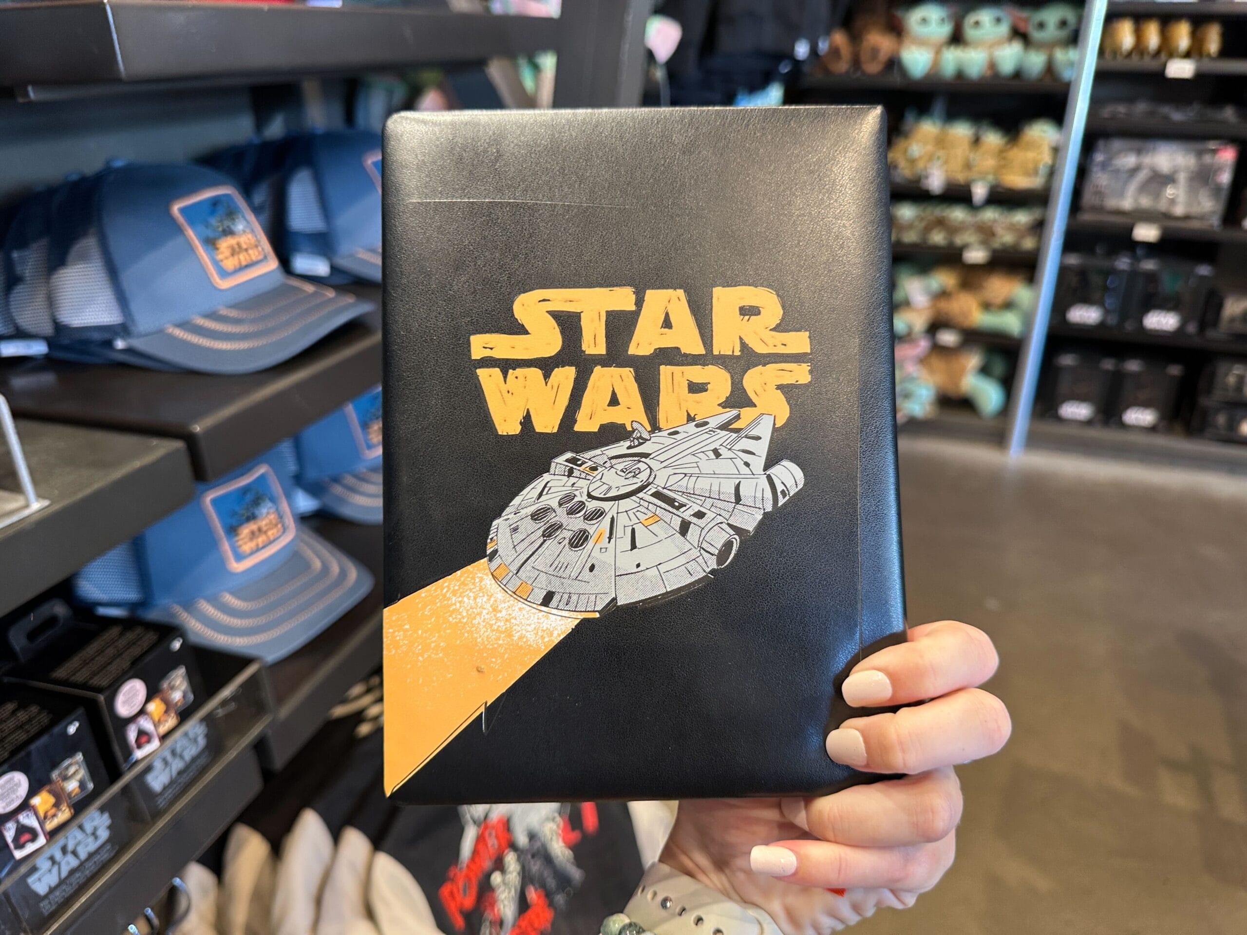Hand holding a black Star Wars Millennium Falcon notebook in a Disney park shop with themed apparel and merch in the background.