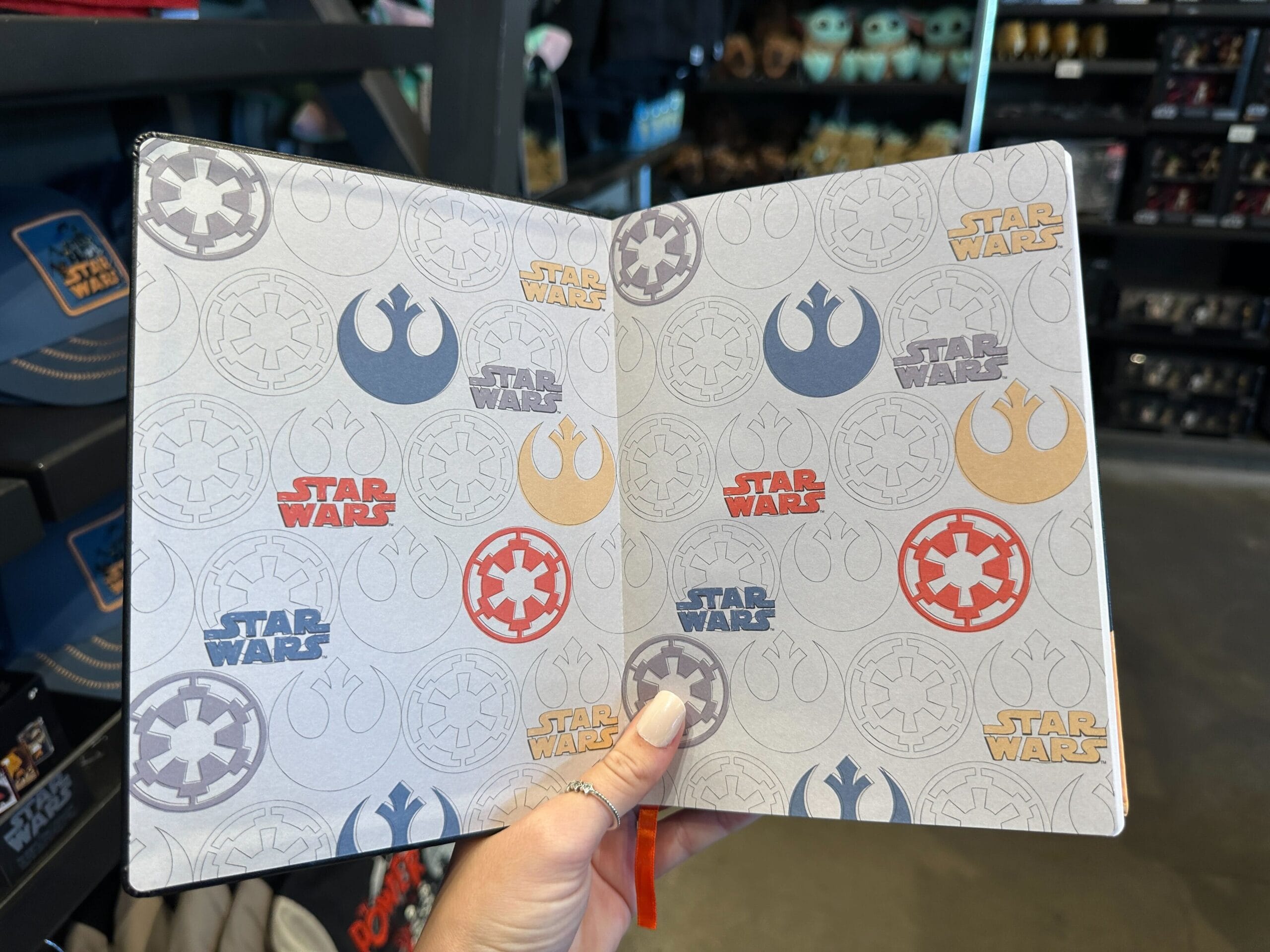 A hand displays a Star Wars-themed notebook among themed apparel inside a Disney World shop, with iconic park merchandise visible nearby.