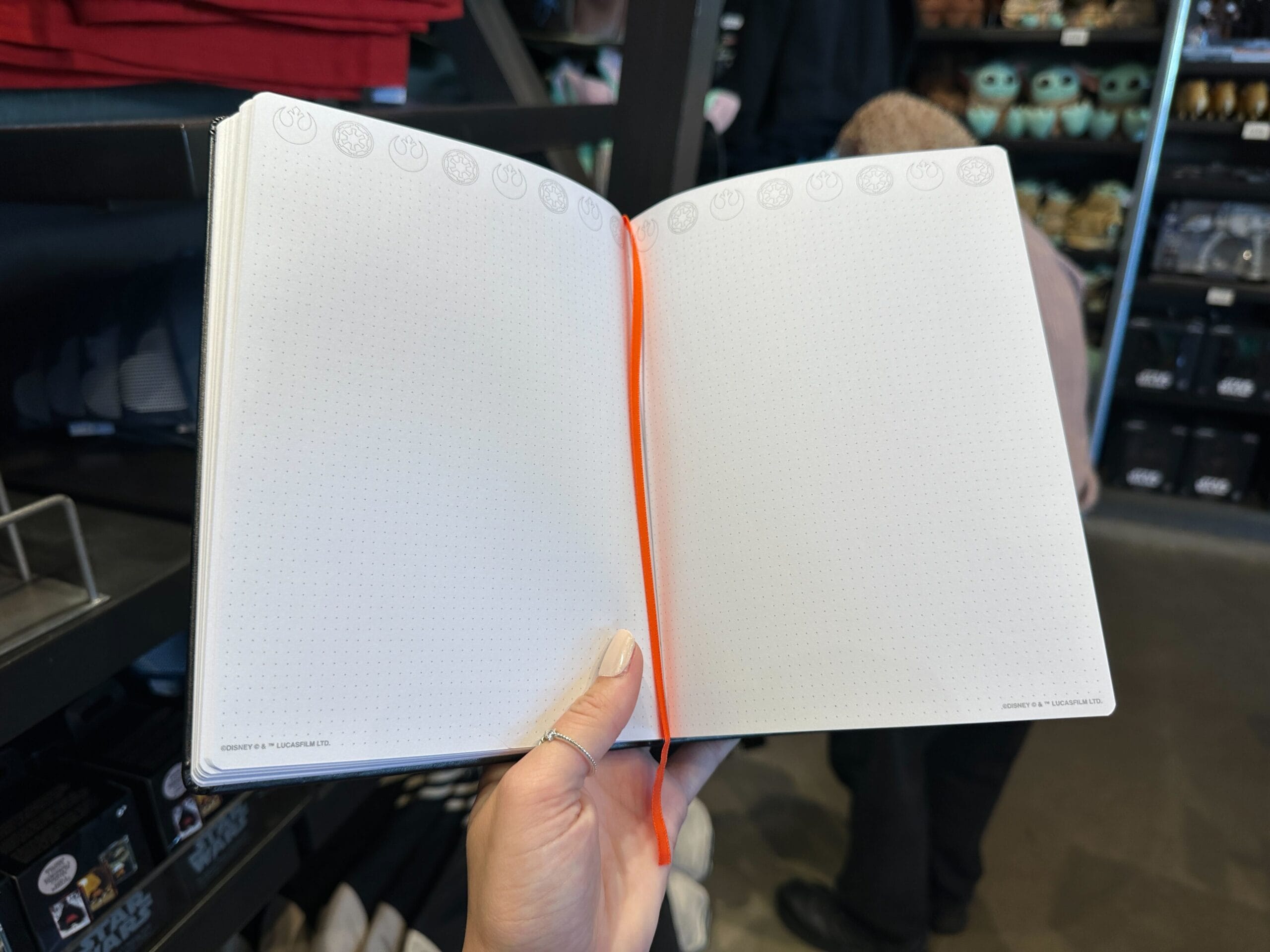 A hand holds open a blank dotted Star Wars notebook among Star Wars clothes, much like Star Wars shops at Disney Parks globally.