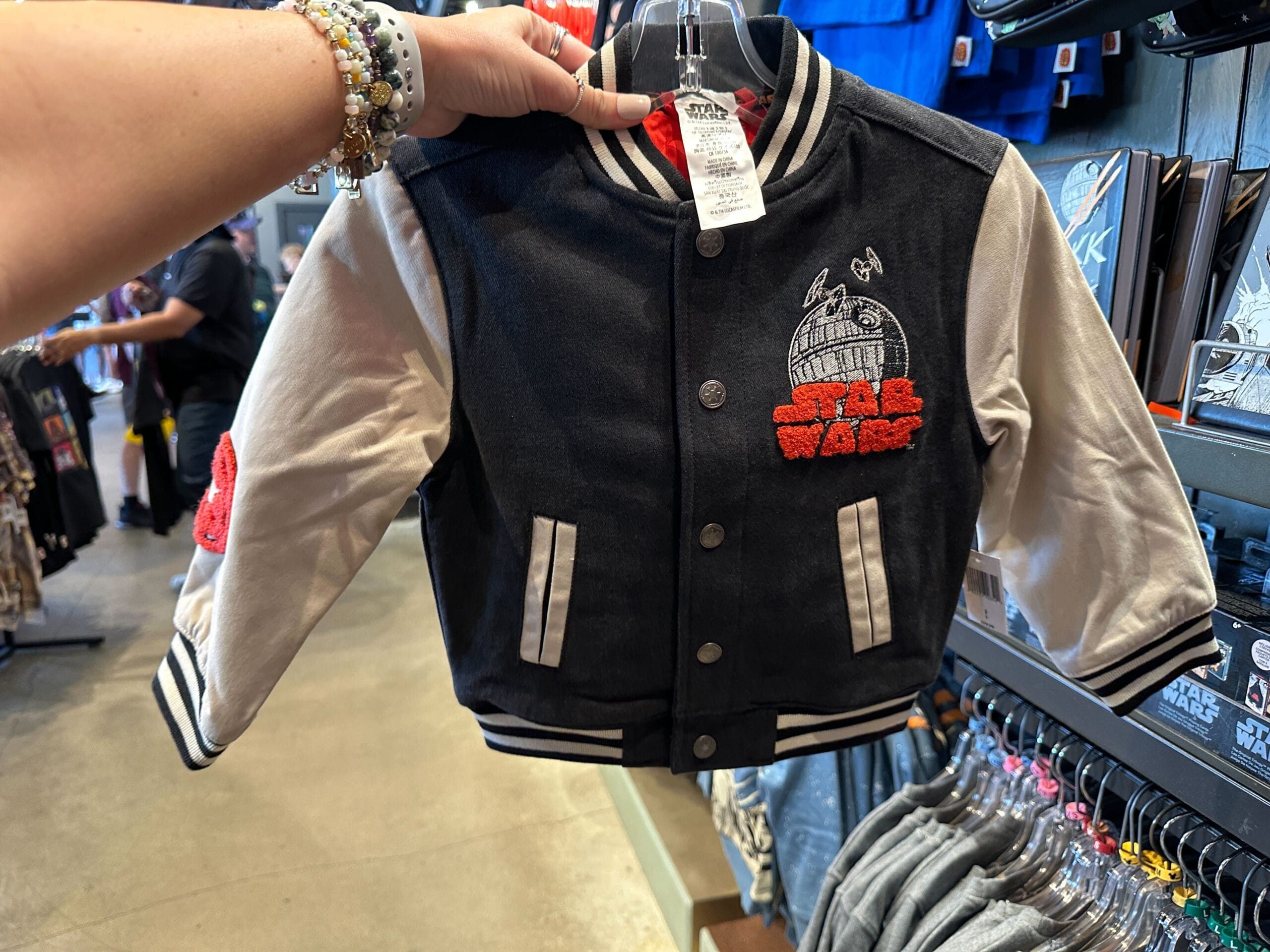 Person holds a toddler's Star Wars varsity jacket with Death Star patch among Star Wars apparel at a Disney park store.
