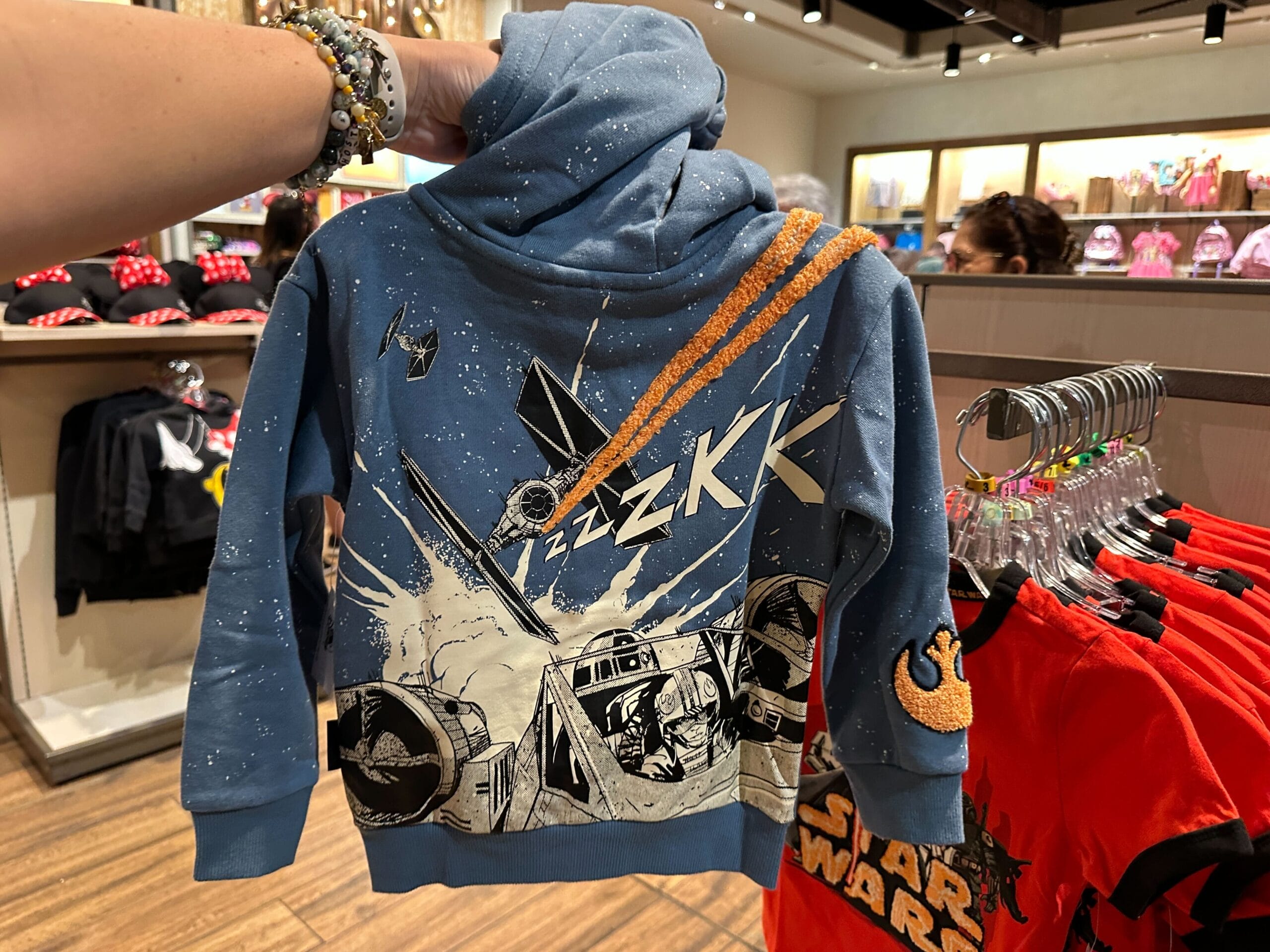 A person holds a blue Star Wars hoodie with a TIE fighter graphic in a Disney park shop, surrounded by park merchandise displays.