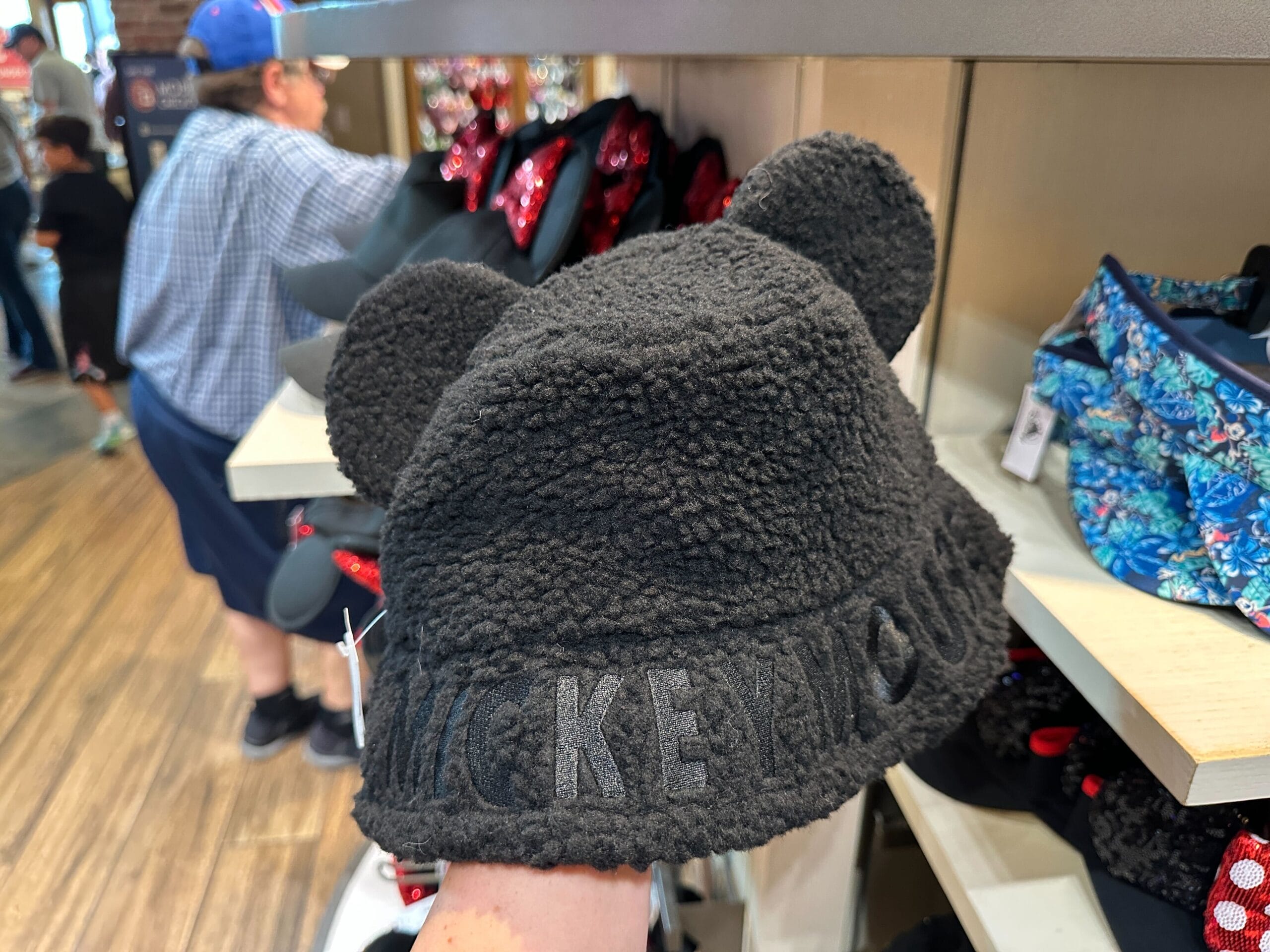 A hand displays a fuzzy Mickey Mouse Sherpa Hat with bear ears and "MICKEY MOUSE" stitched, ideal for chilly Disney park days.