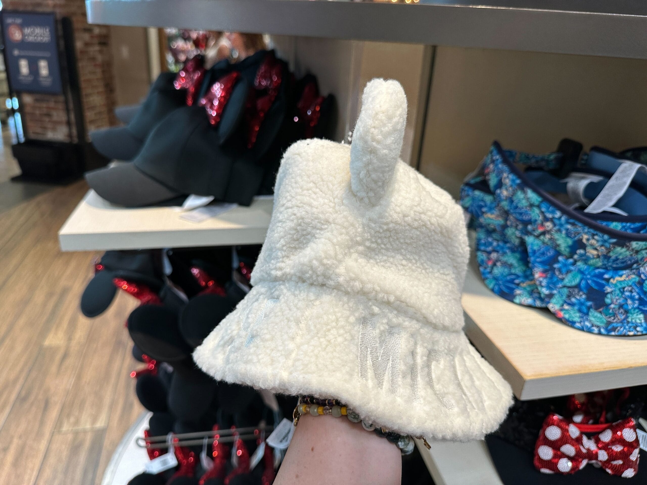A hand displays a fuzzy Mickey Mouse Sherpa Hat with "MICKEY" as Minnie ear hats fill Disney World shelves in the background.