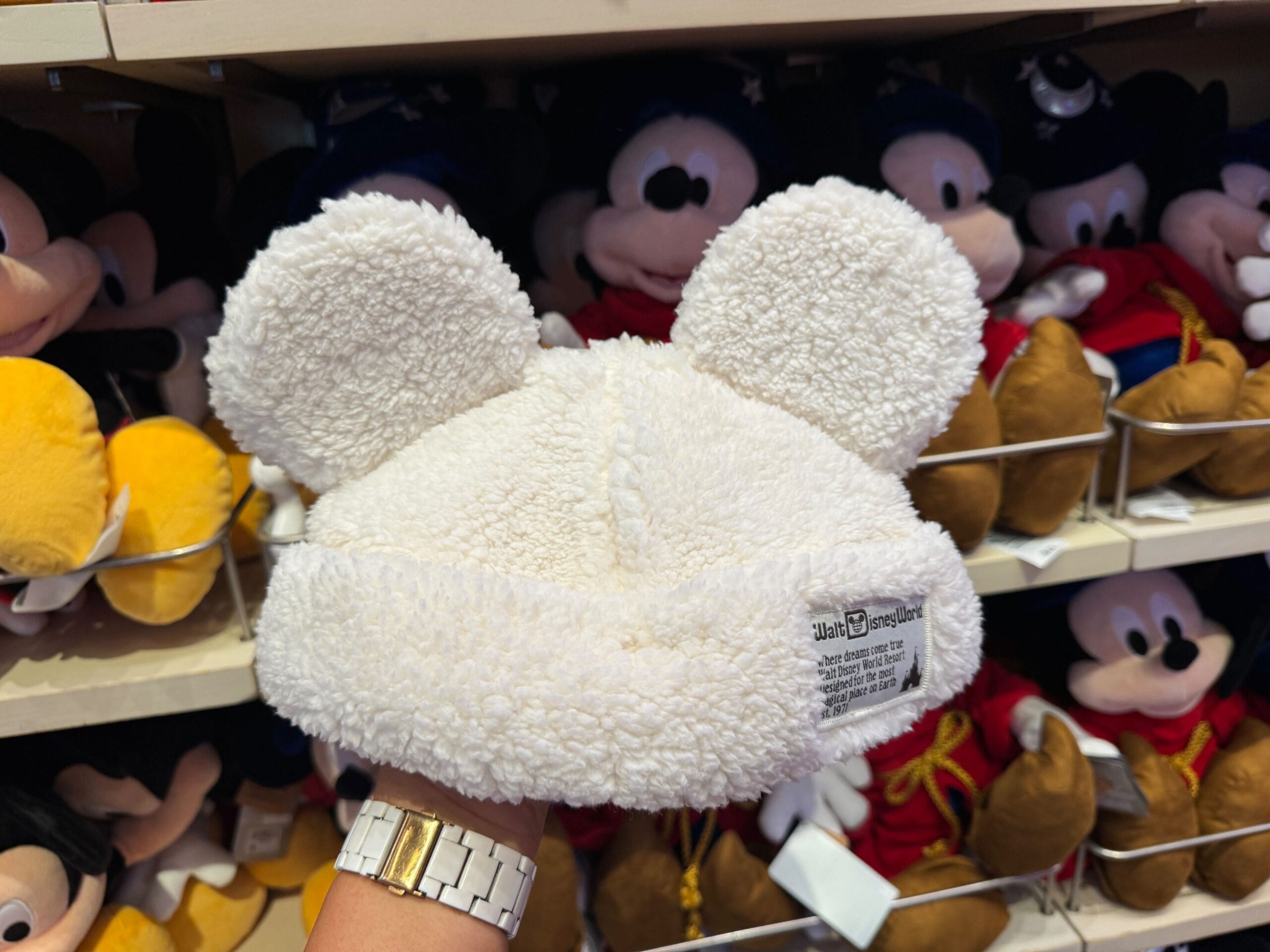 A hand holds a fluffy white winter cap with bear ears in front of shelves filled with Mickey Mouse plush toys.