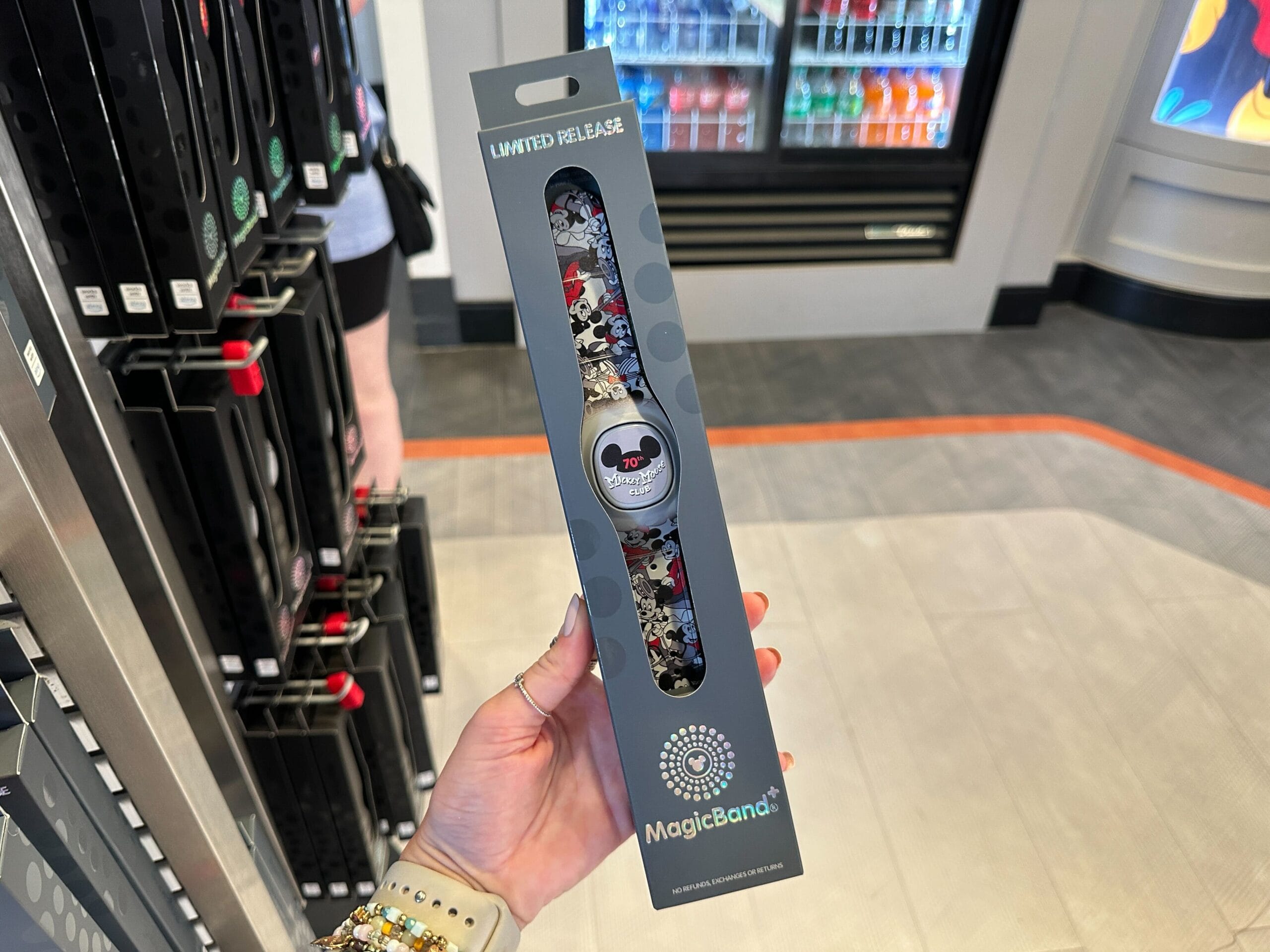 Person holding a Mickey Mouse Club MagicBand+ in front of a store display and drink cooler.