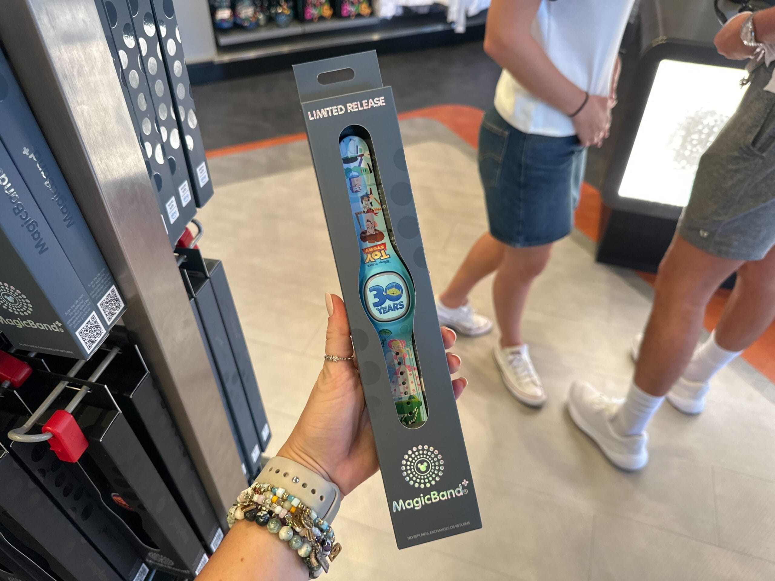 Hand holding a limited release MagicBand+ in store, with people and MagicBand displays in the background.