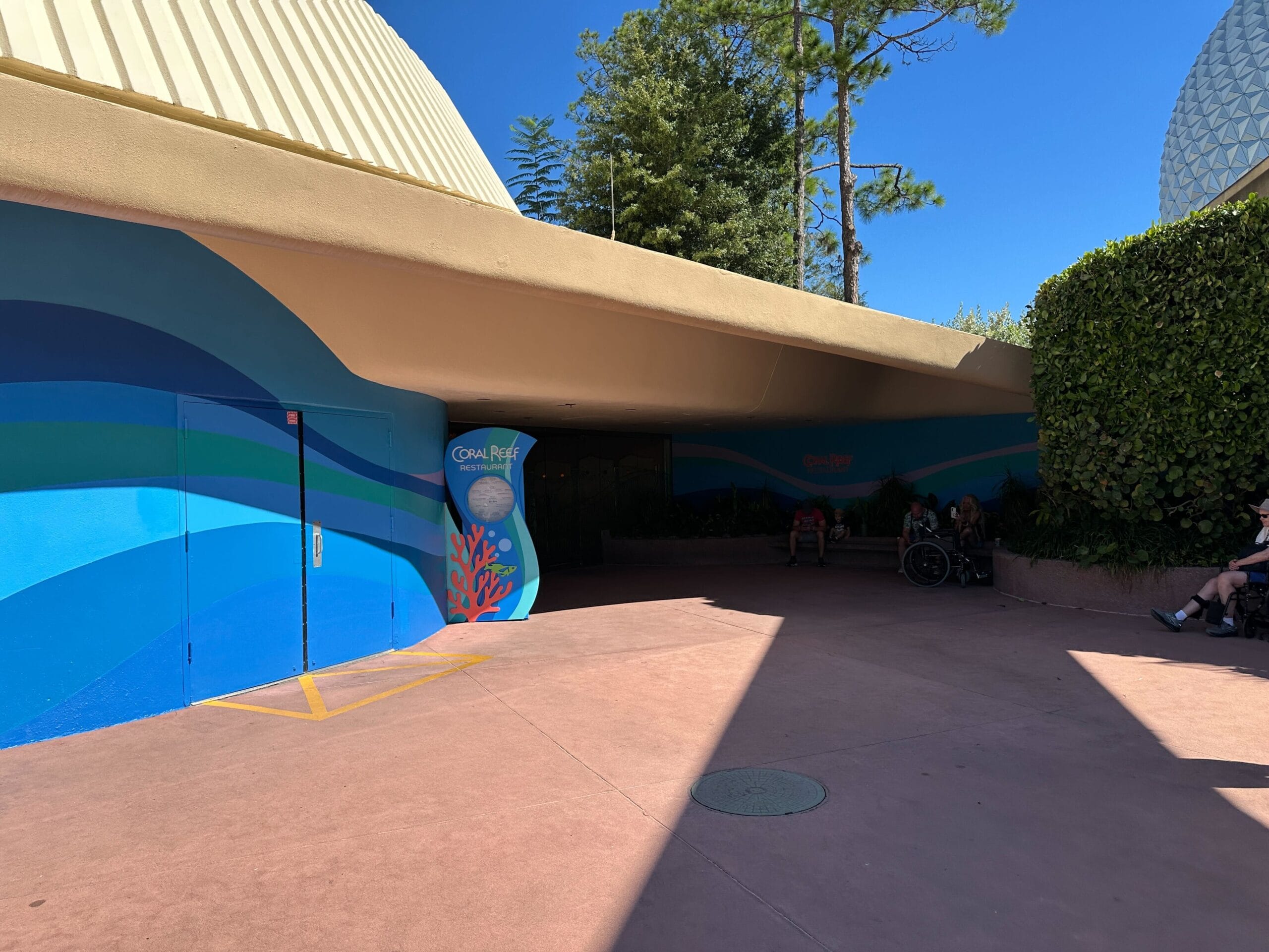 Building entrance with a blue wave mural, round sign, and shaded spot, reminiscent of classic 1980s EPCOT Center design at Disney.