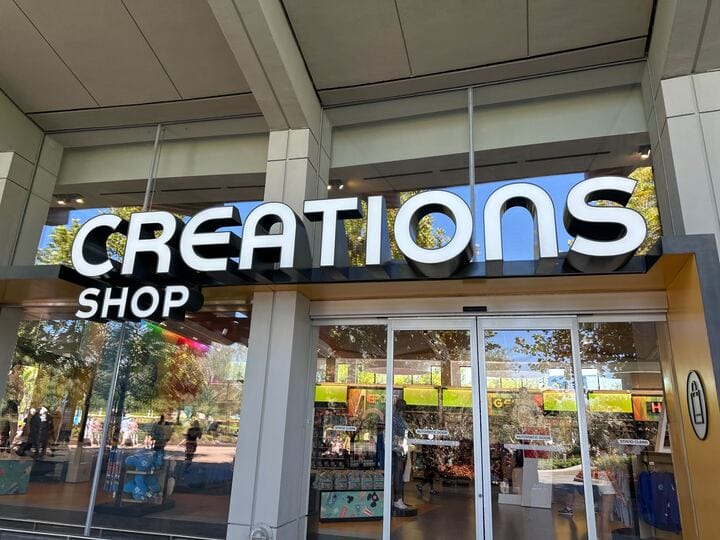 Creations Shop storefront at EPCOT with bold white sign, trees and sky reflected—discover Authentic Original Jackets unique to Disney.
