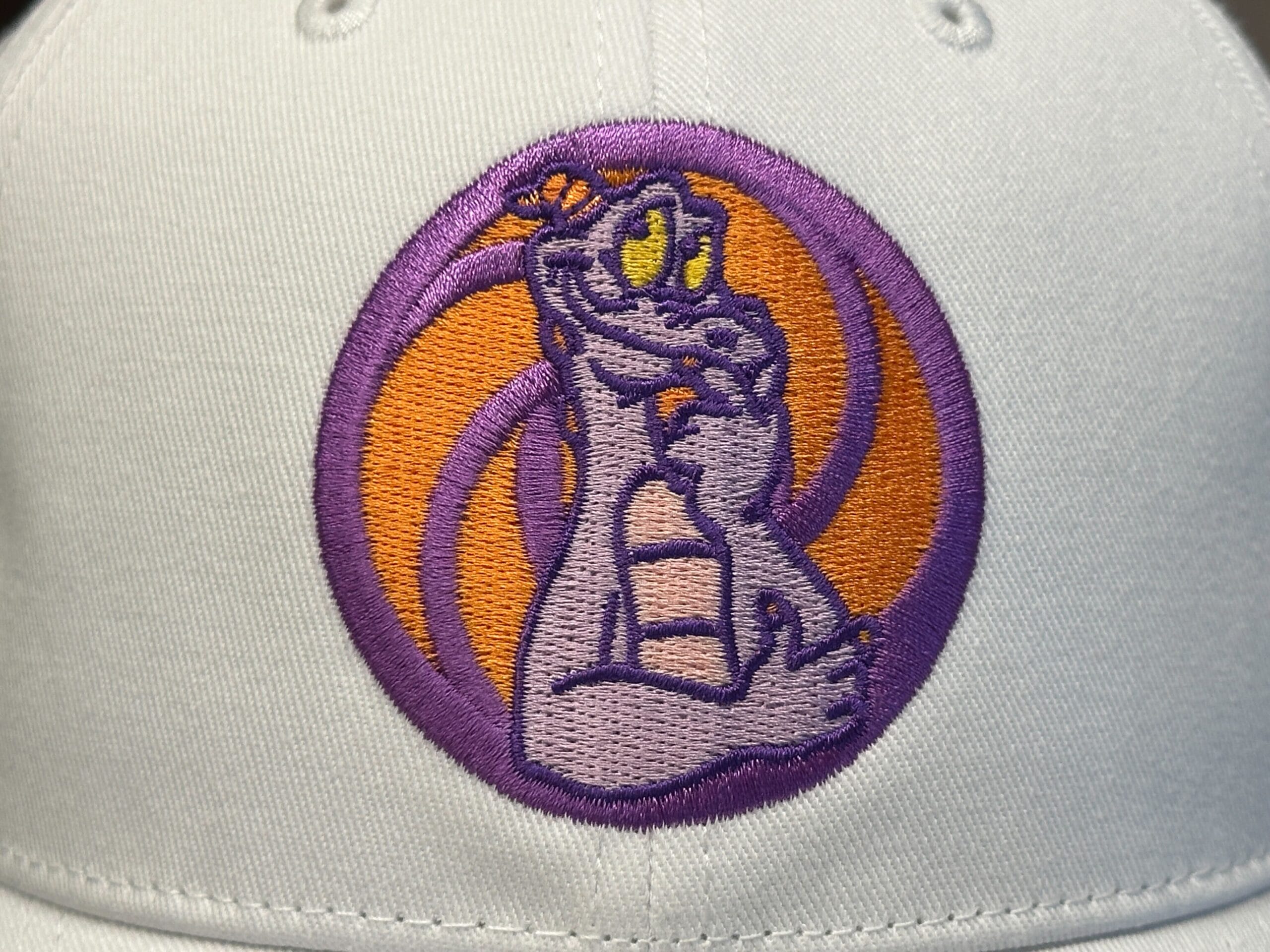 Embroidered cartoon purple dragon with yellow eyes on a white Figment Baseball Cap, Disney World-inspired background.