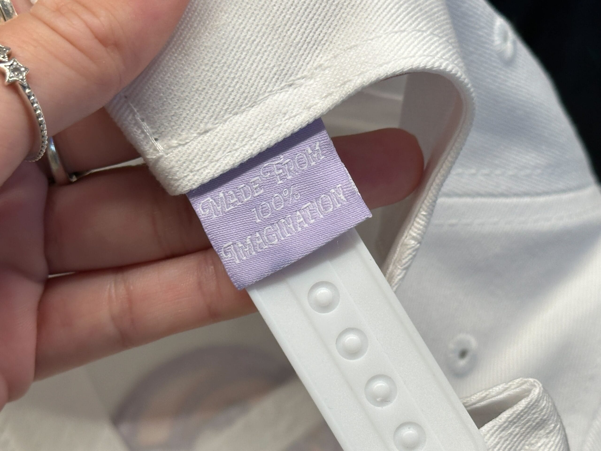 A hand holds a white Figment Baseball Cap with a lavender tag that reads "MADE FROM 100% IMAGINATION.