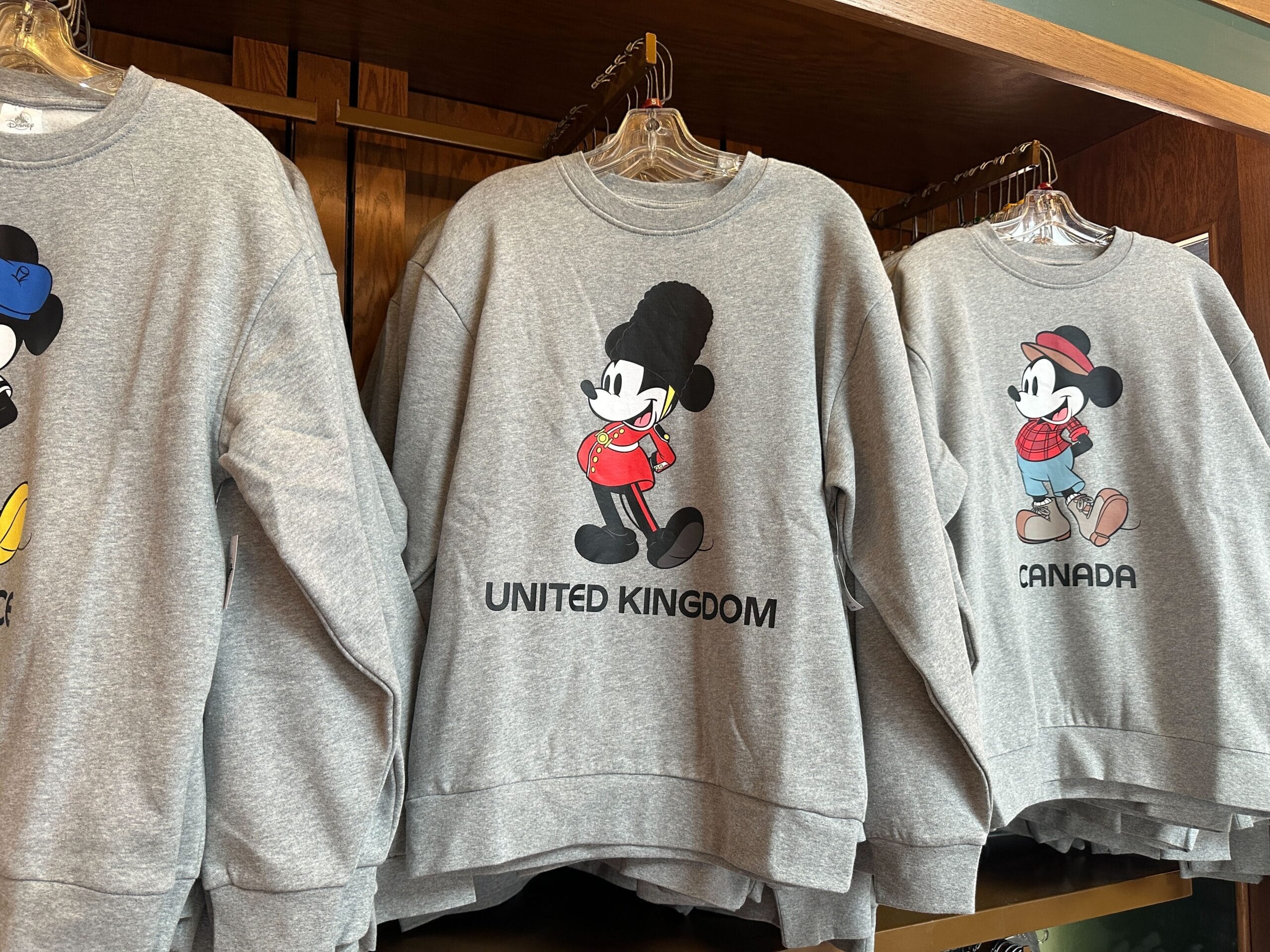 Mickey Mouse sweatshirts from the EPCOT Sweatshirt Collection showcase UK and Canada outfits on a rack.
