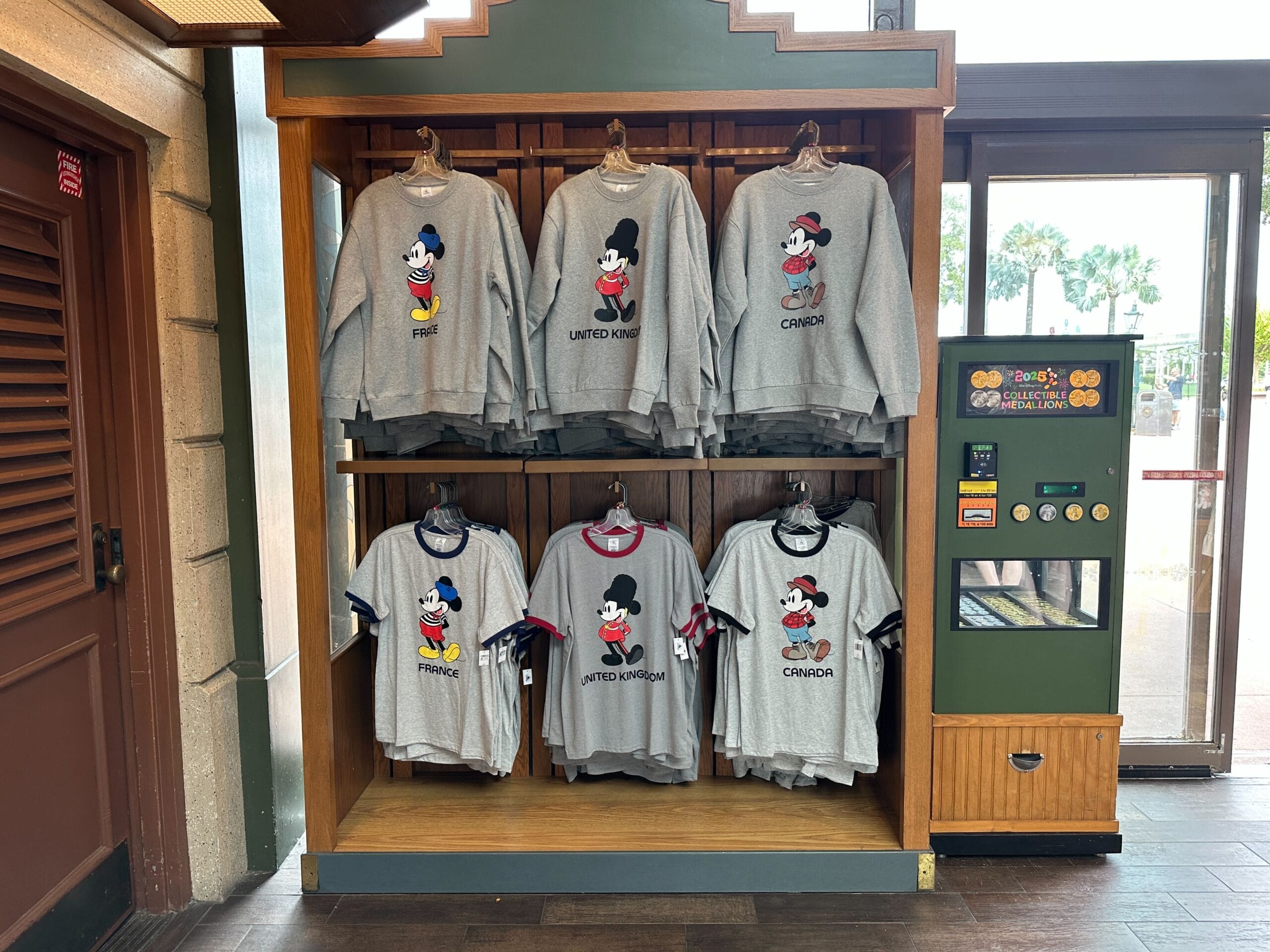 The EPCOT Sweatshirt Collection features gray Mickey Mouse sweatshirts labeled France, UK, and Canada on a wooden shelf.