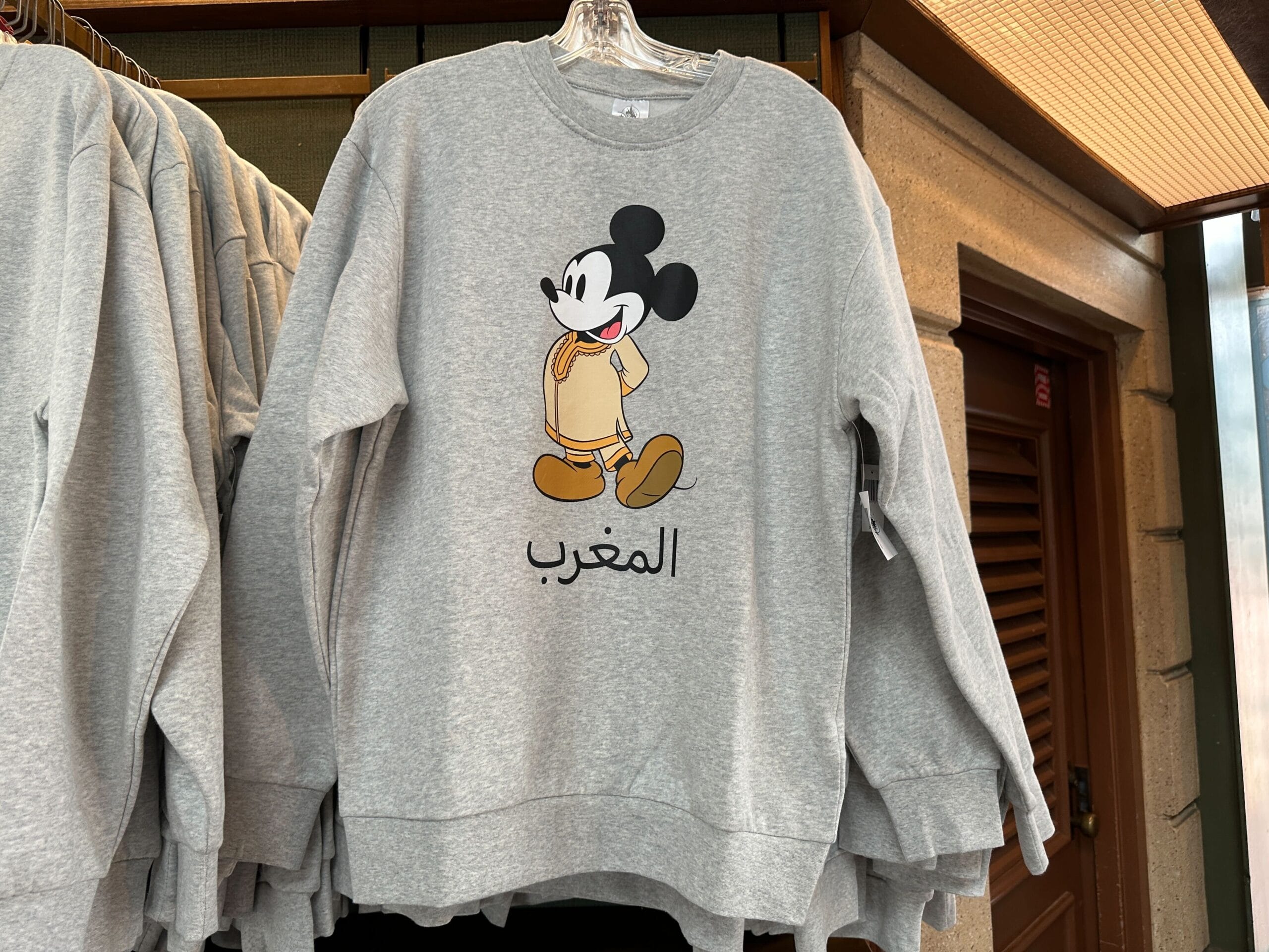 Gray World Showcase Sweatshirt with Mickey Mouse and Arabic "Morocco" text, displayed on a hanger in a shop.